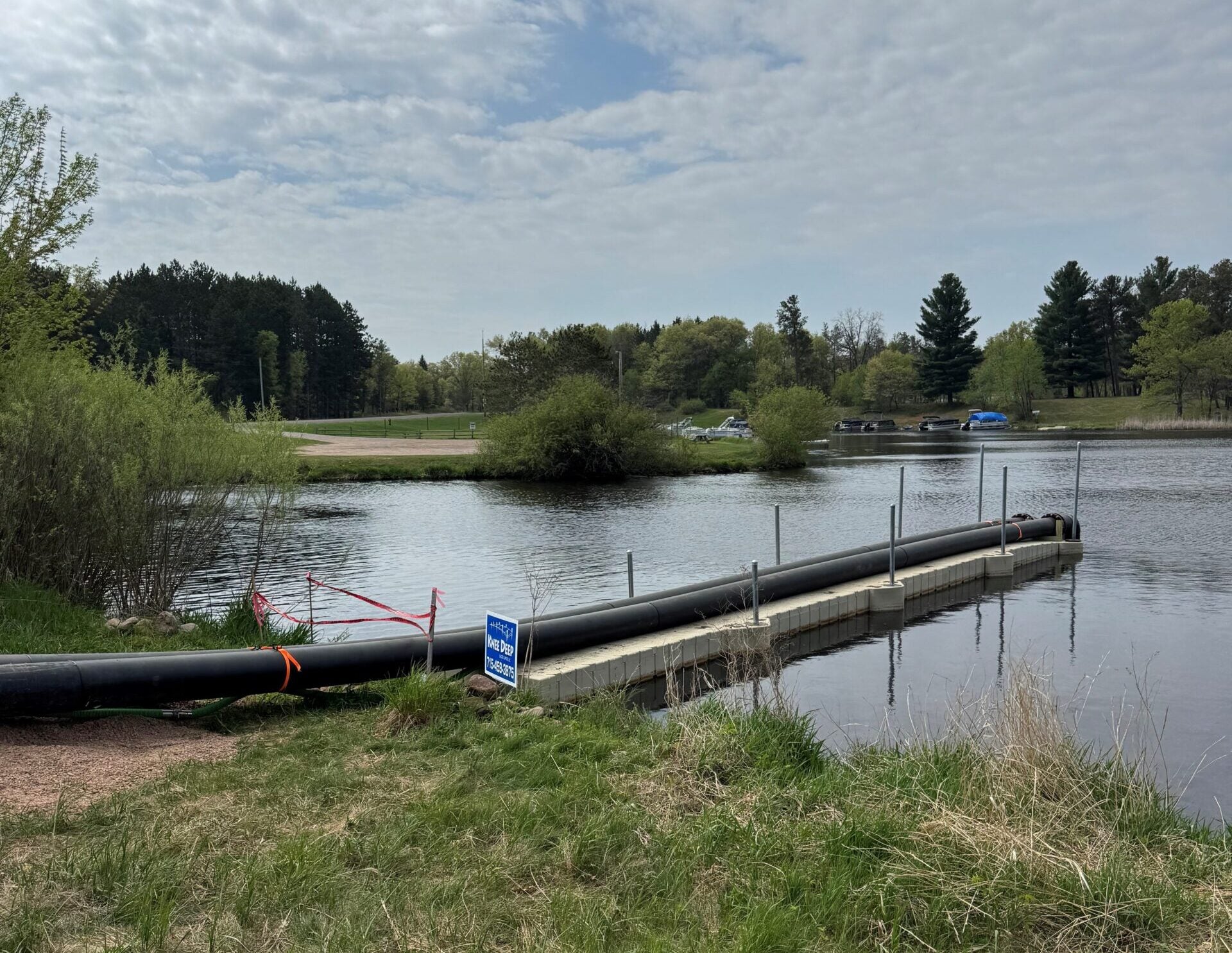 A floating dock extends into a calm lake under a cloudy sky, with a restricted access sign and grassy shoreline in the foreground. Trees and parked cars are visible in the background.