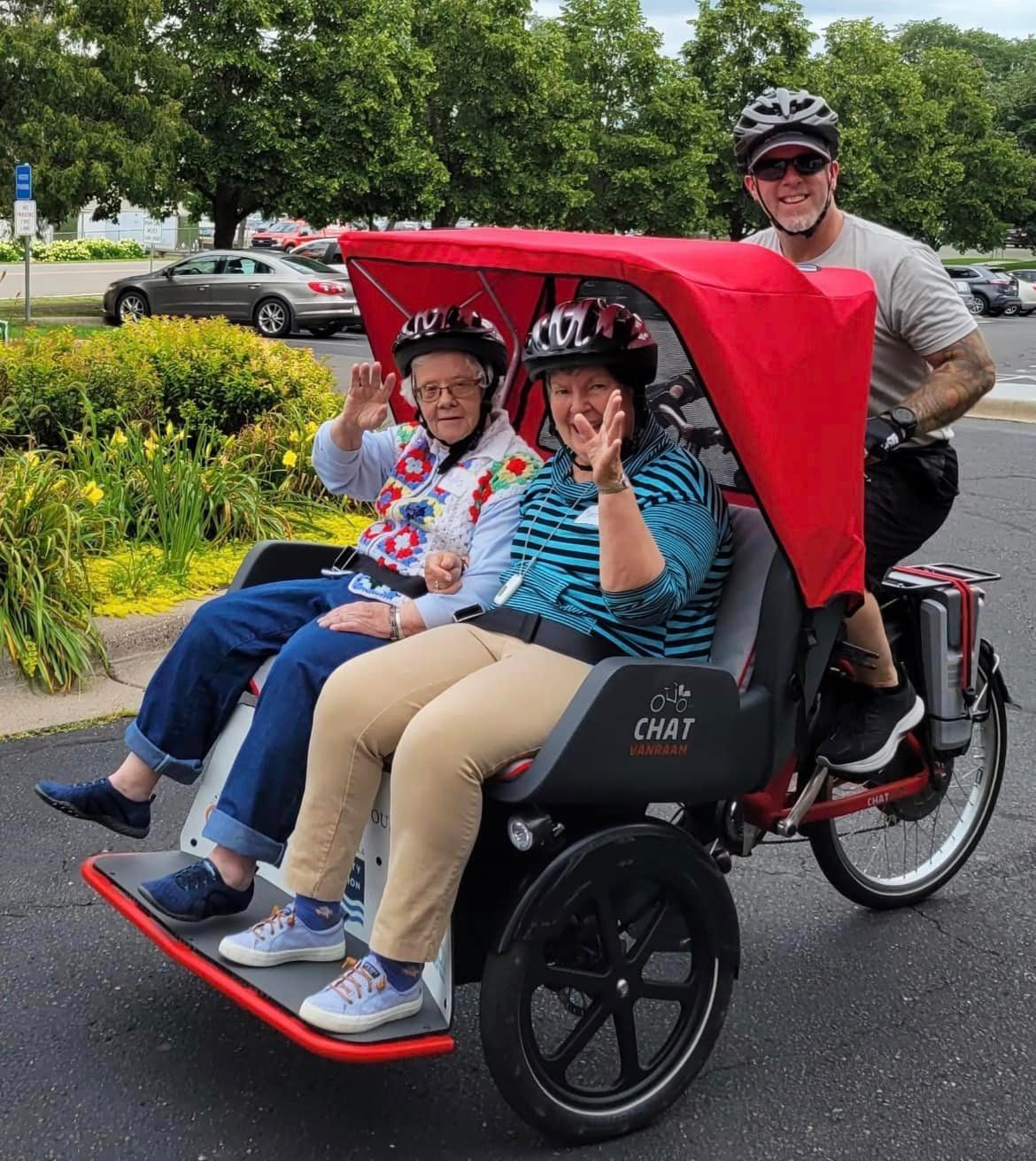 Two older women wearing helmets ride in a cycle rickshaw with a red canopy, driven by a man in a helmet, all smiling and waving on a paved path with greenery in the background.