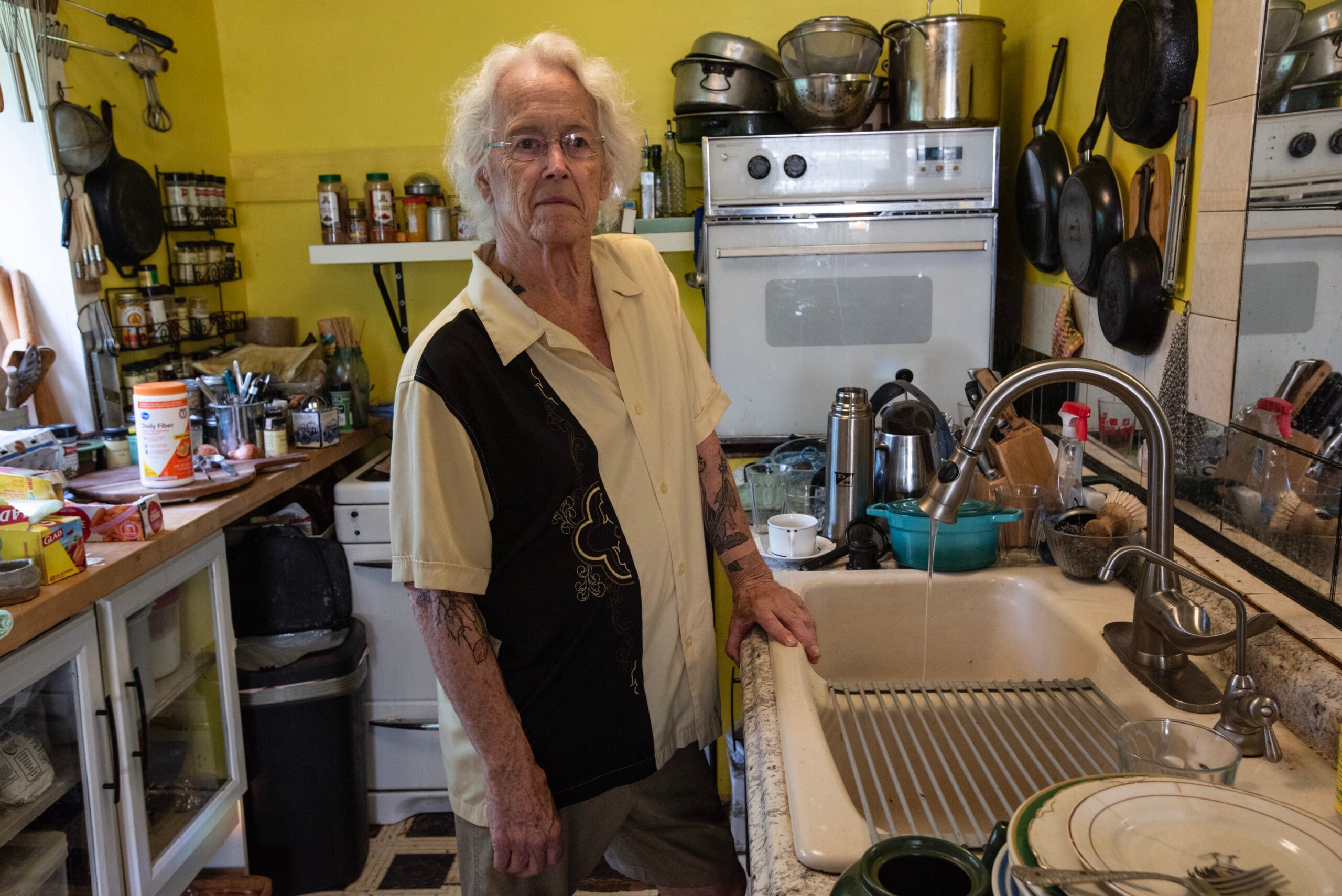 An older person with white hair stands by a kitchen sink, surrounded by dishes and cooking utensils, in a small, cluttered kitchen with yellow walls.