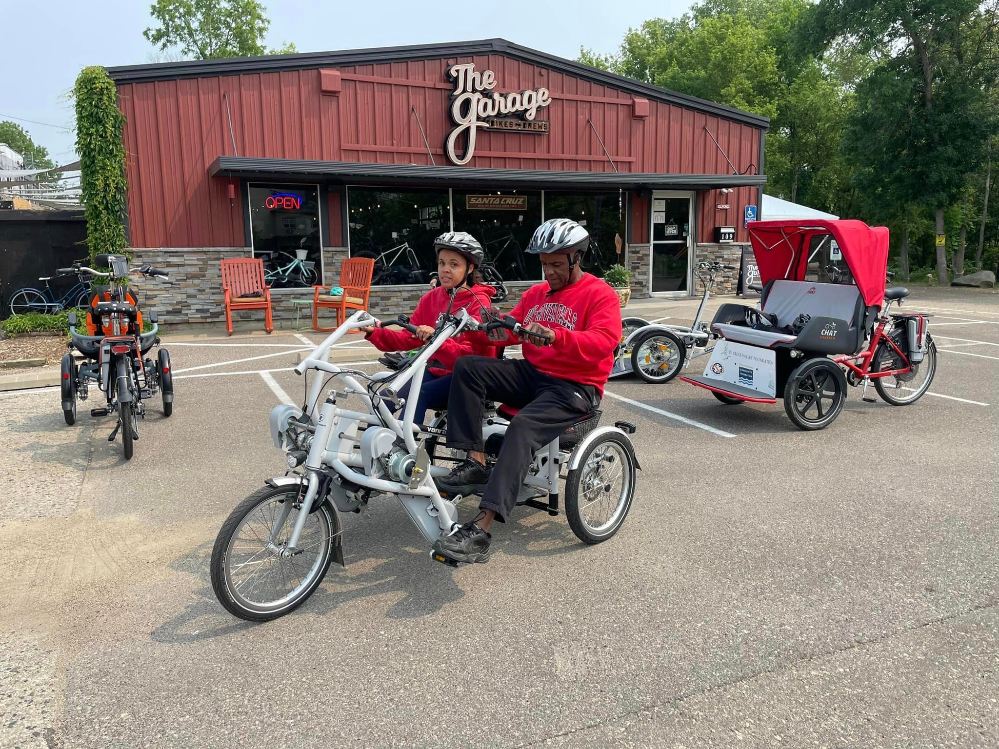 Two people ride a tandem adaptive tricycle in front of a building called “The Garage,” with other adaptive bikes and a rickshaw-style trike visible in the parking lot.