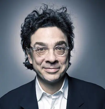 Man with curly dark hair, glasses, and a suit jacket over a white shirt, smiling at the camera, with a gradient grey background.