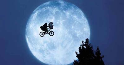 A silhouette of two figures on a bicycle flies in front of a large full moon against a night sky, with tree tops visible below.