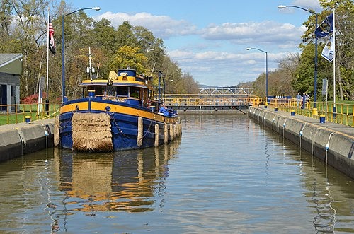 A blue and yellow boat is positioned inside a canal lock with gates at both ends, surrounded by trees and a bridge visible in the background.