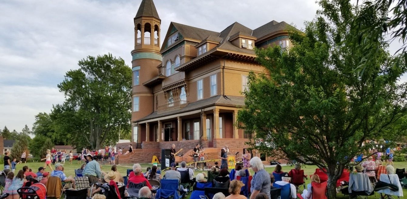 A crowd sits on lawn chairs and blankets in front of a large, historic, Victorian-style building during an outdoor event.