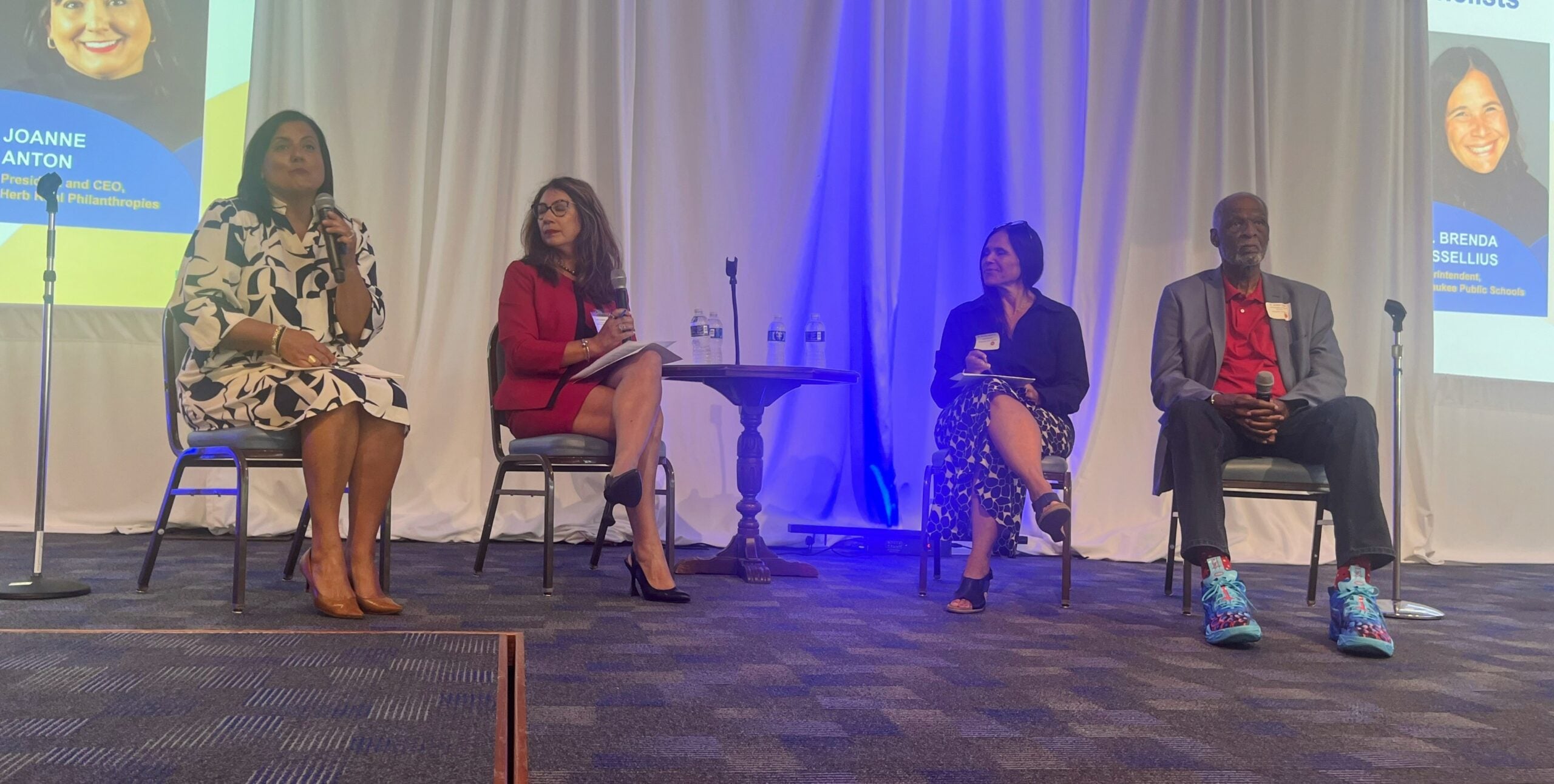 Four panelists, three women and one man, sit on chairs on a stage with a white curtain backdrop during a discussion event. Two water bottles are on a small table between them.