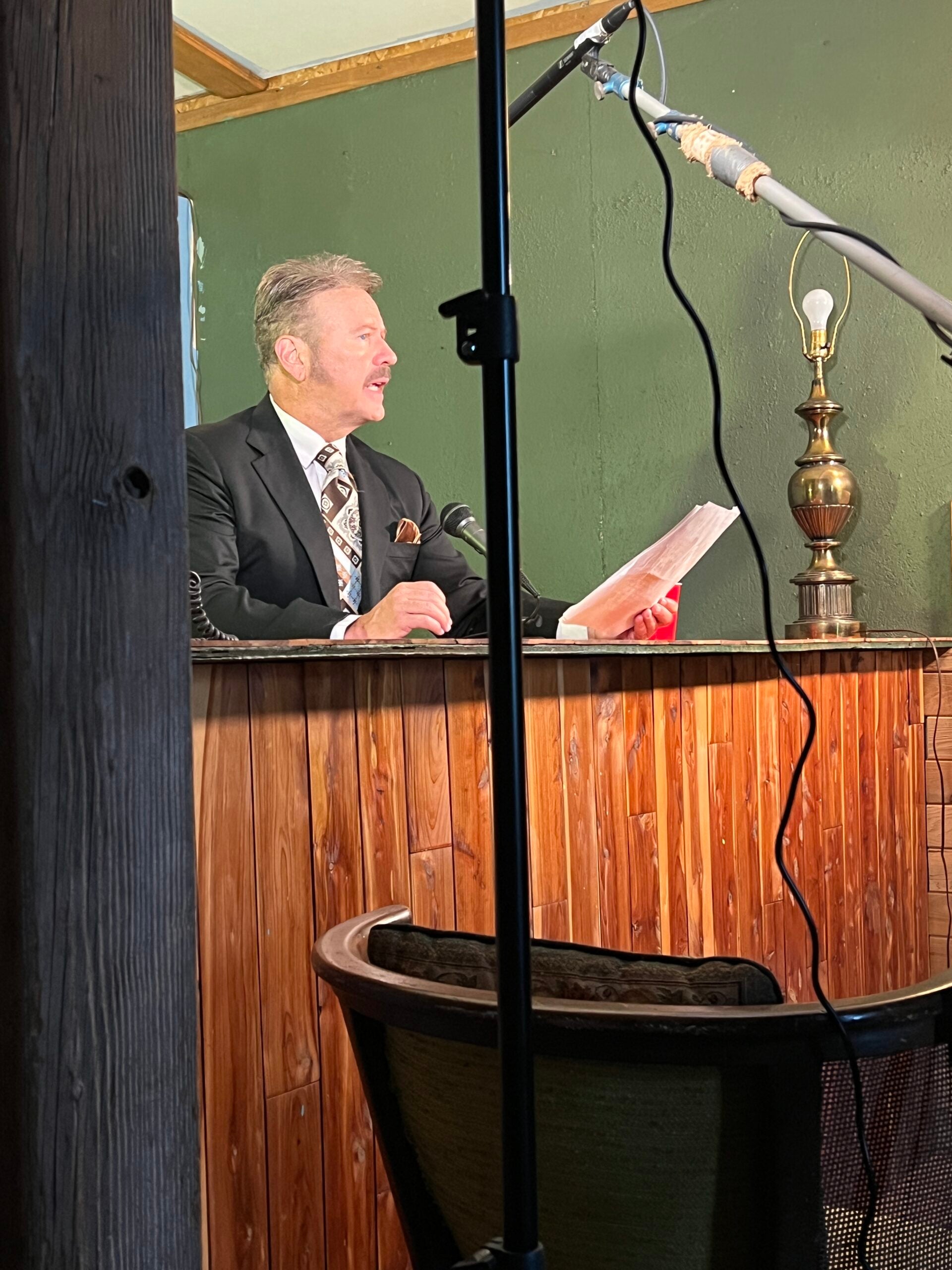 A man in a suit sits at a wooden desk holding papers, speaking into a microphone. Studio lights and equipment are visible around him.