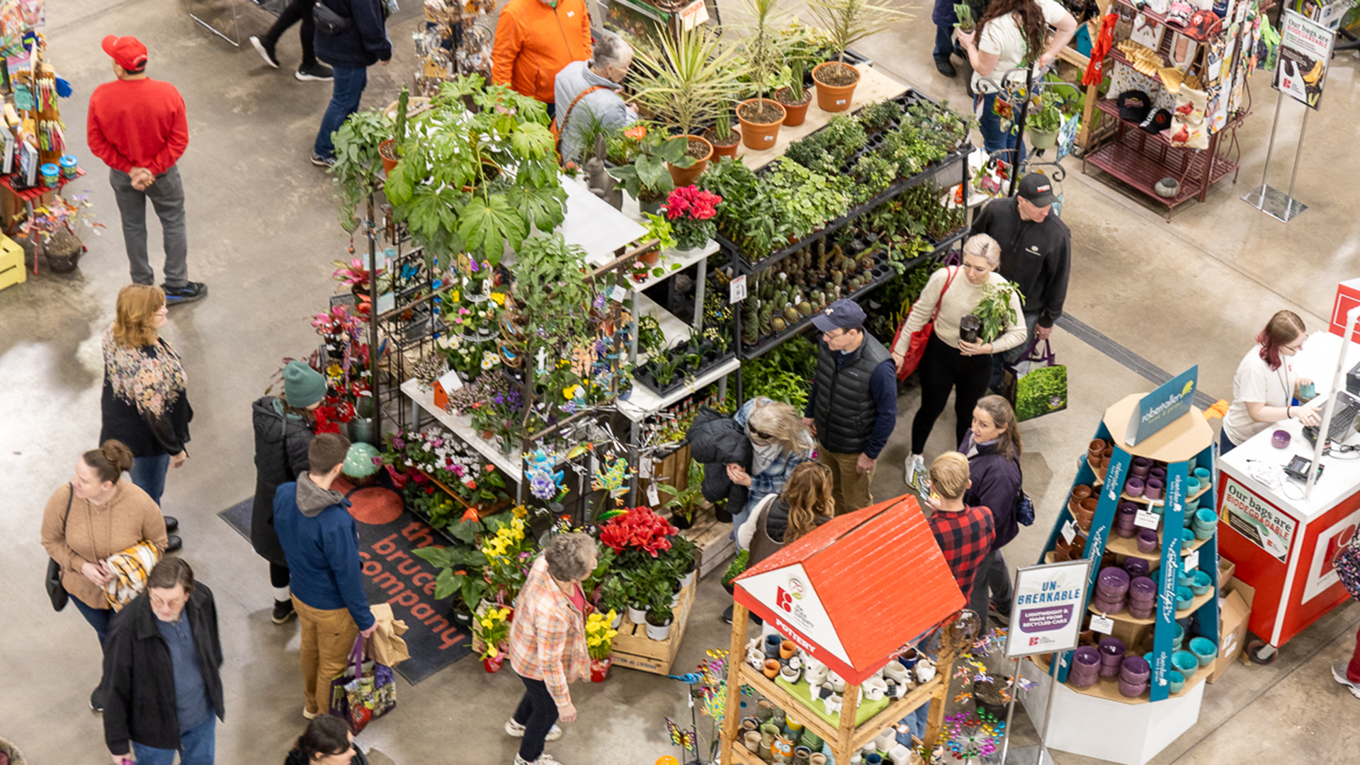 People shop for plants and garden supplies at a busy indoor market, with various plants and gardening items displayed on tables and shelves.