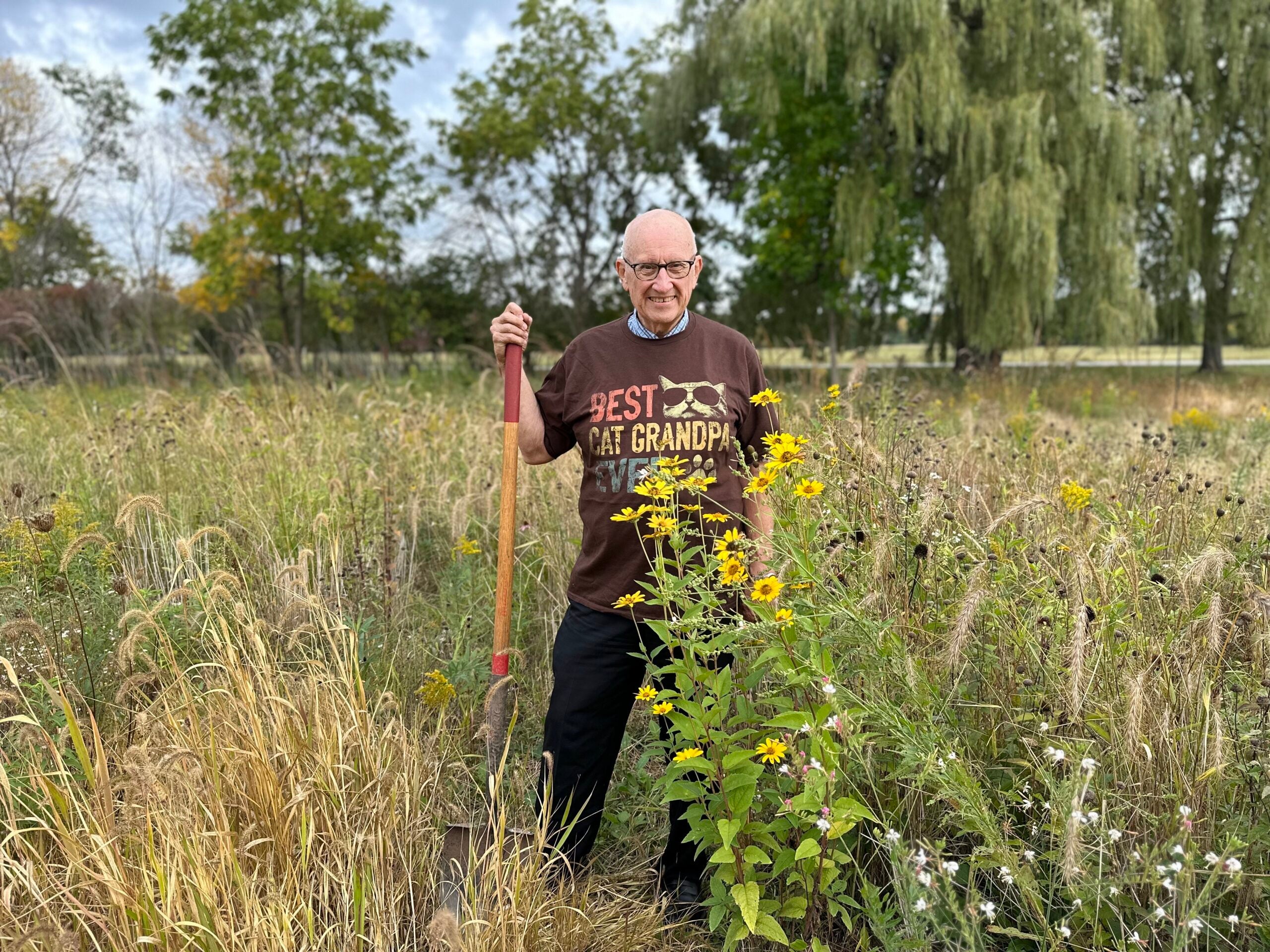 An older man stands in a grassy field holding a shovel. He wears glasses and a Best Cat Grandpa t-shirt. Yellow wildflowers and tall grasses surround him. Trees are in the background.