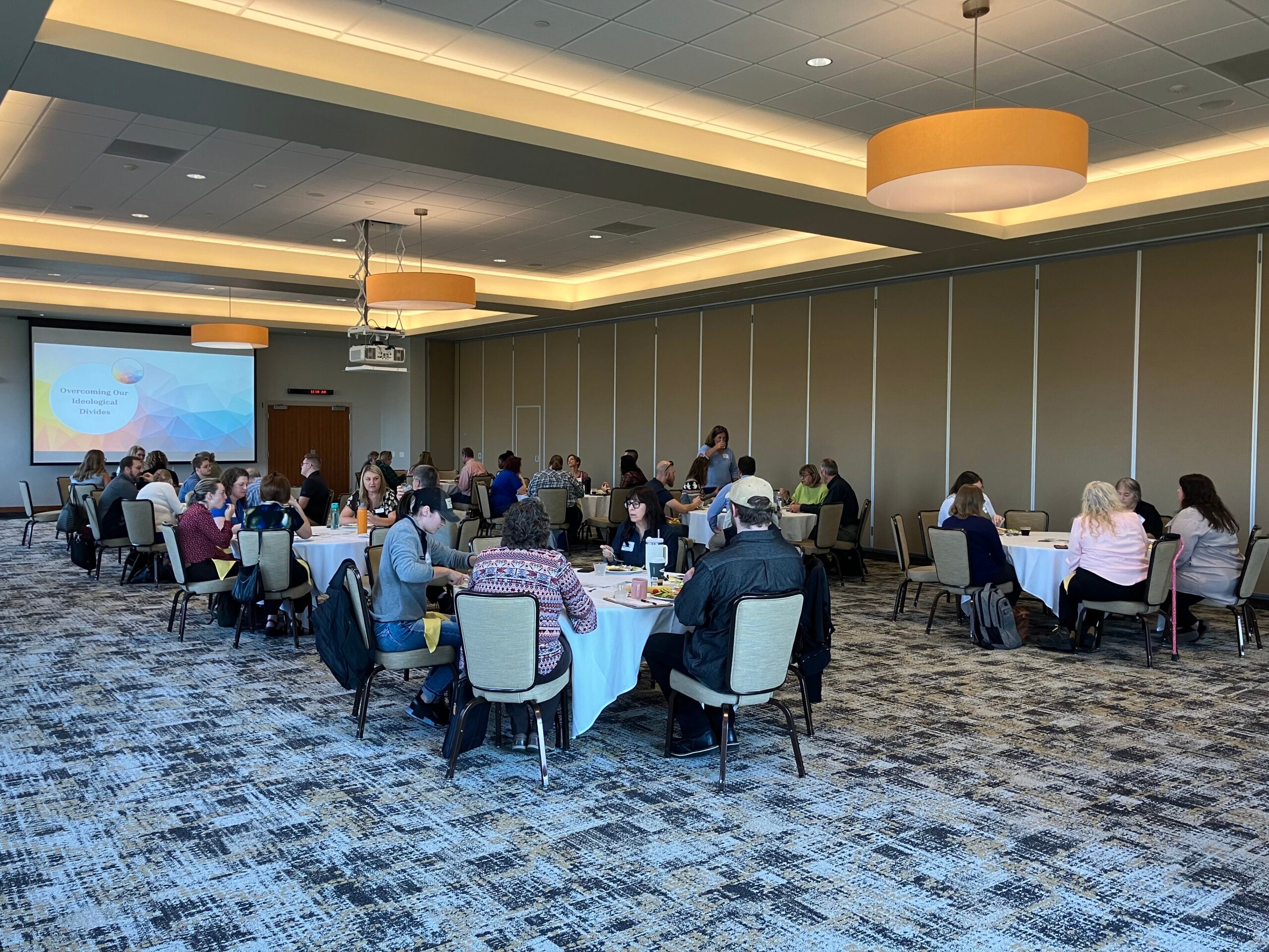 People seated at round tables in a conference room are participating in a group event or workshop. A presentation is displayed on a screen at the front of the room.