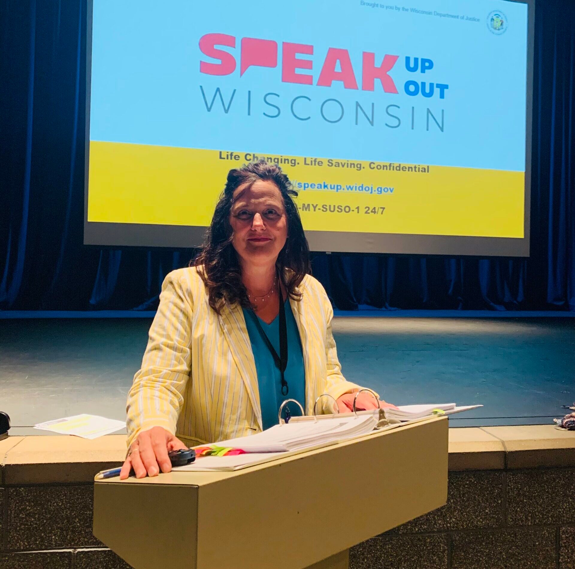 A woman stands at a podium with papers in front of a screen displaying Speak Up Speak Out Wisconsin in a dimly lit auditorium.