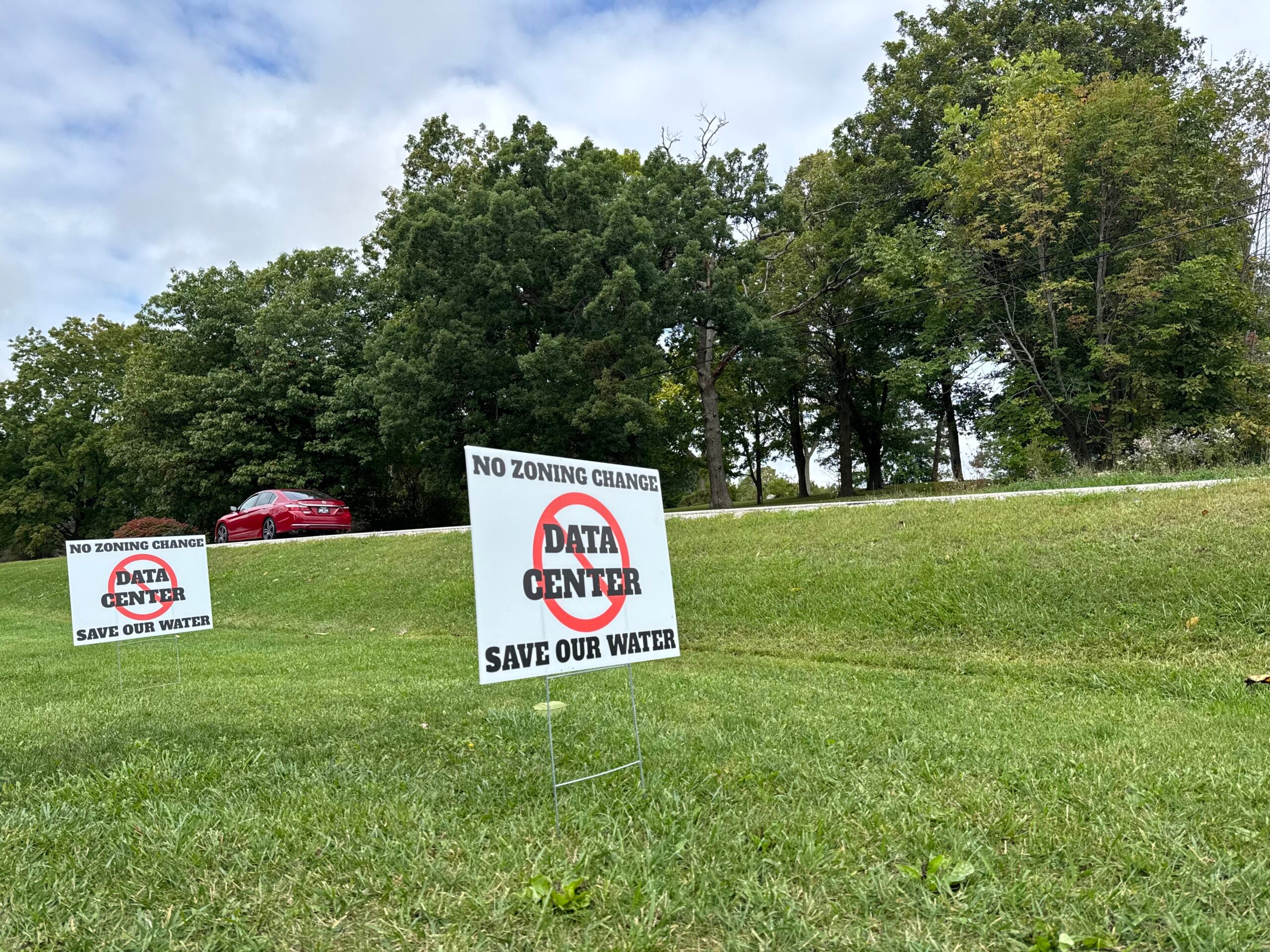 Lawn signs read “No zoning change, No data center, Save our water” near a green grassy area with trees and a red car in the background.