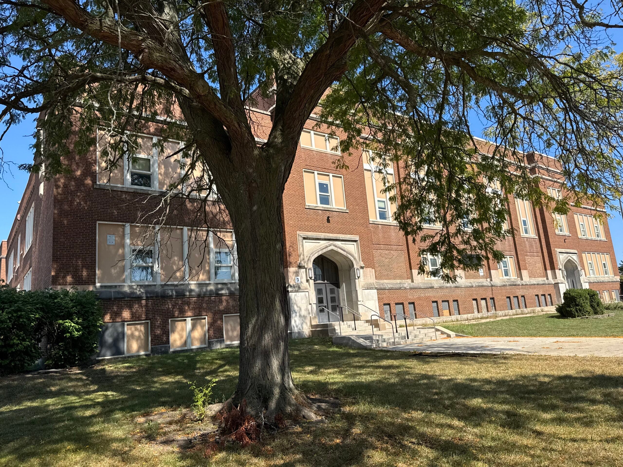A large tree stands in front of a three-story red brick building with arched entrances and several windows on a sunny day.