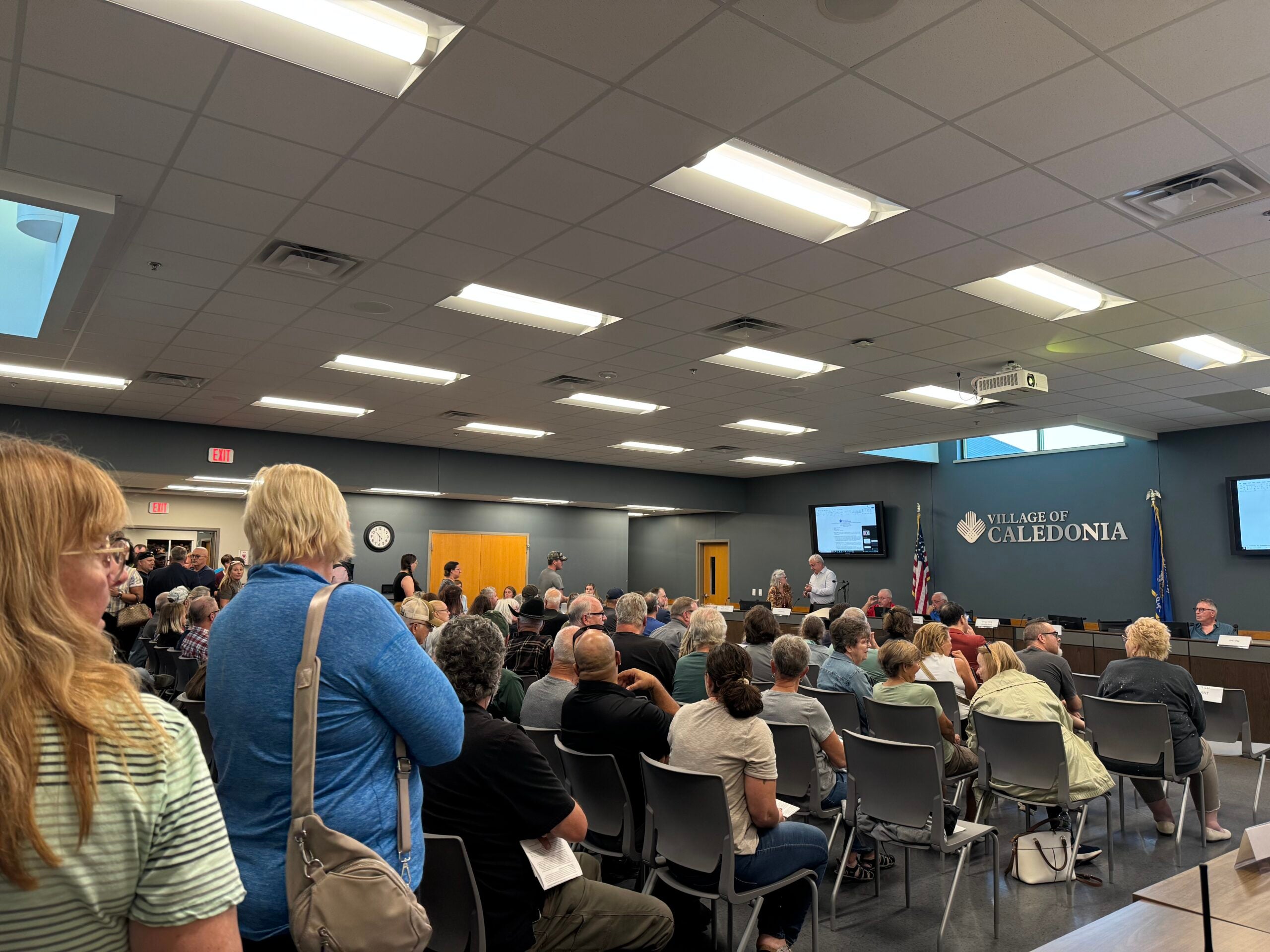 A large group of people seated and standing in a meeting room, facing a dais with officials under a sign reading Village of Caledonia.