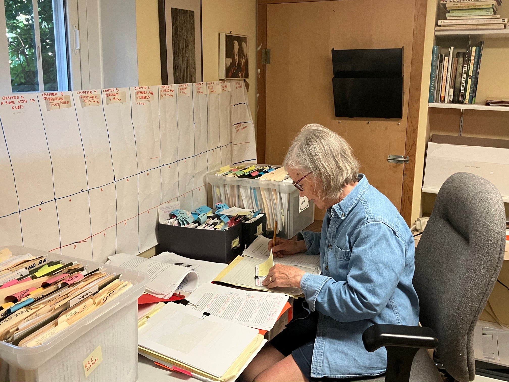 An older woman sits at a desk organizing papers and files, with charts and timelines taped to the wall behind her in a home office setting.
