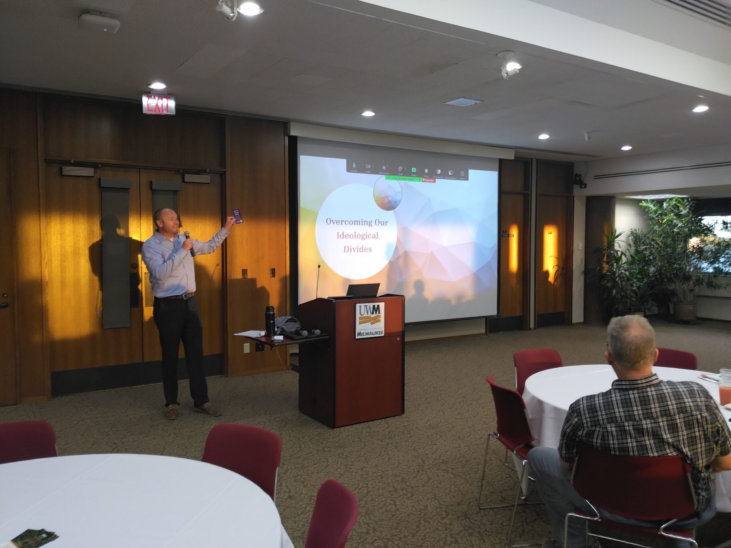A man stands beside a podium presenting a slide titled Overcoming Our Ideological Divides to a small audience in a conference room.
