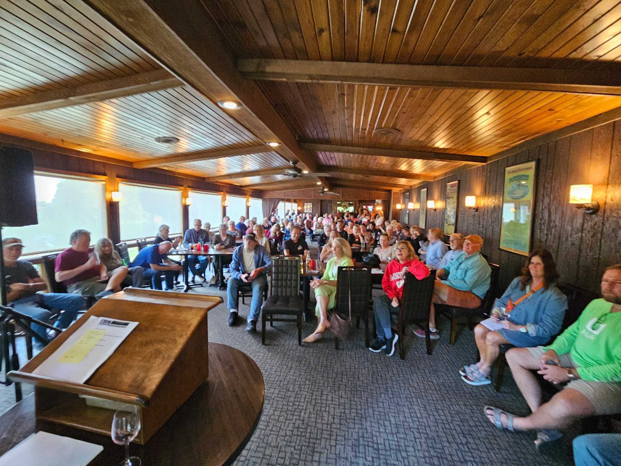 A large group of people sit in a wood-paneled room, facing a podium, suggesting a meeting or event is taking place.
