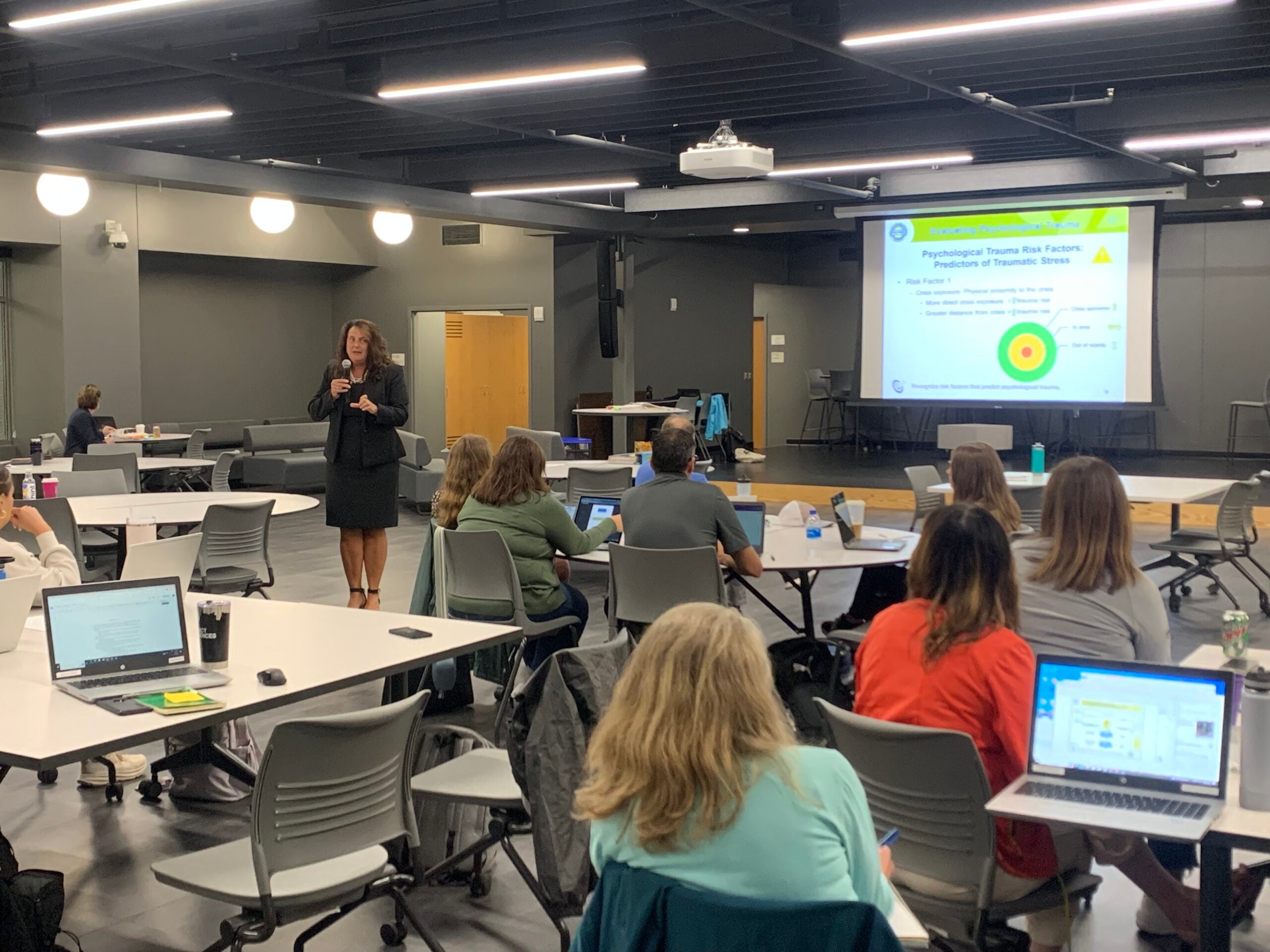 A woman stands and speaks to a group of people seated at tables with laptops in a conference room, while a presentation is projected on a screen.