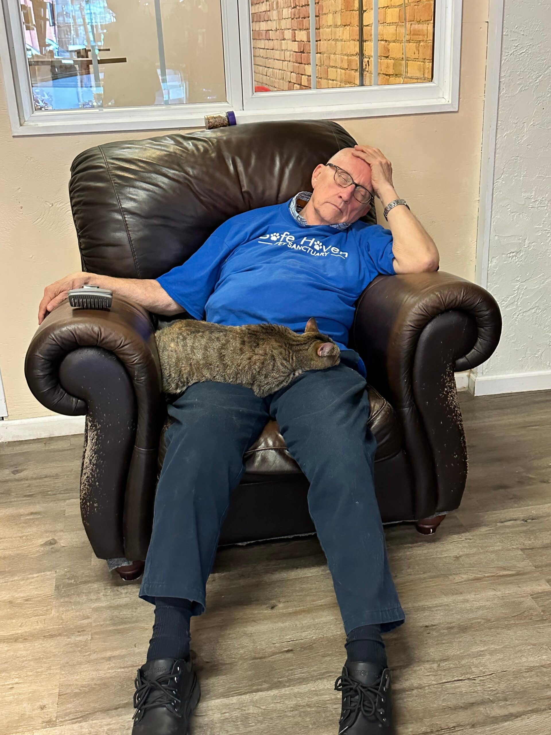 An elderly man sleeping in a brown leather armchair with a remote on the armrest and a tabby cat curled up on his lap.