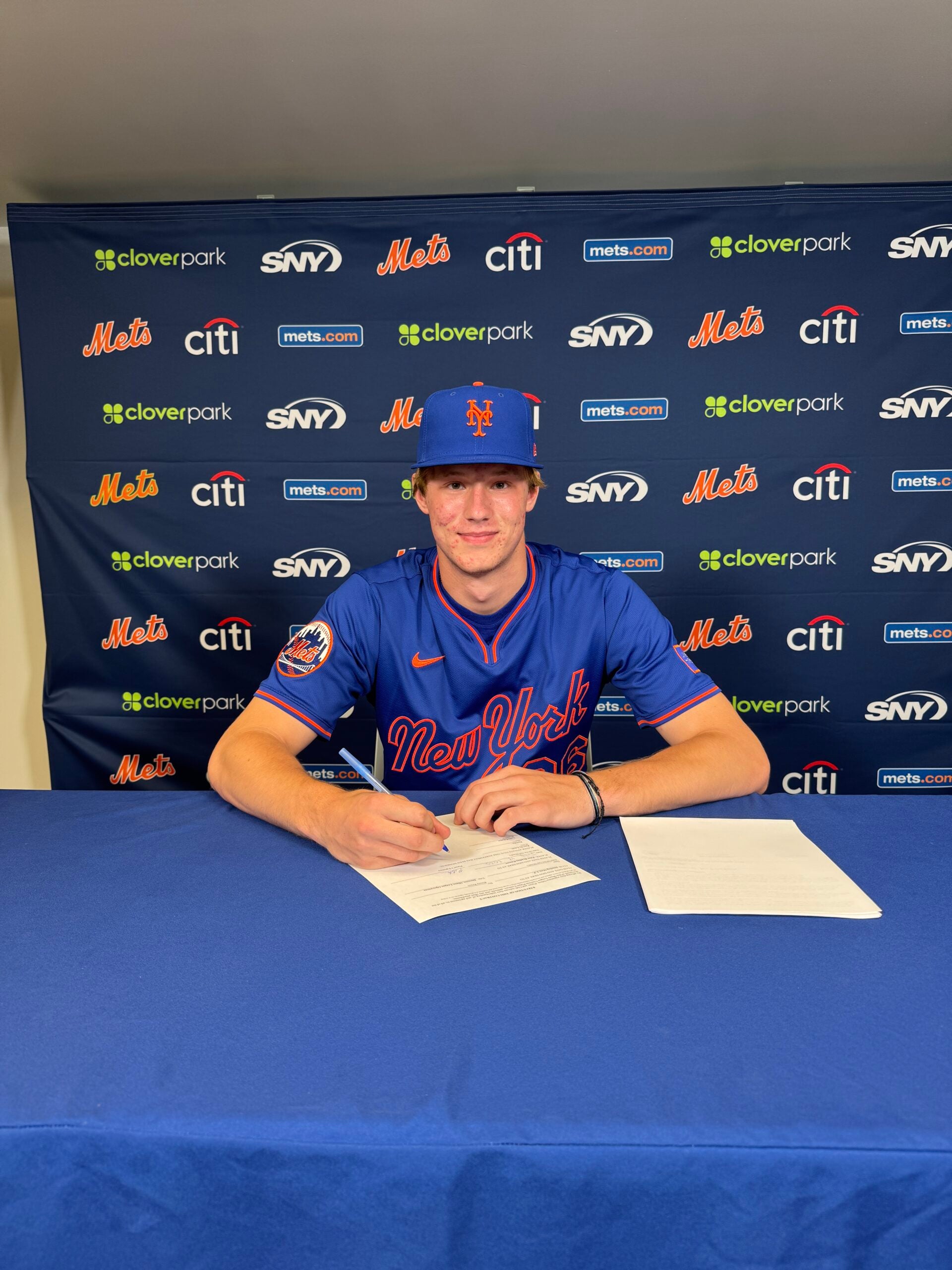 A baseball player in a New York Mets uniform sits at a table signing documents, with a Mets-branded backdrop behind him.