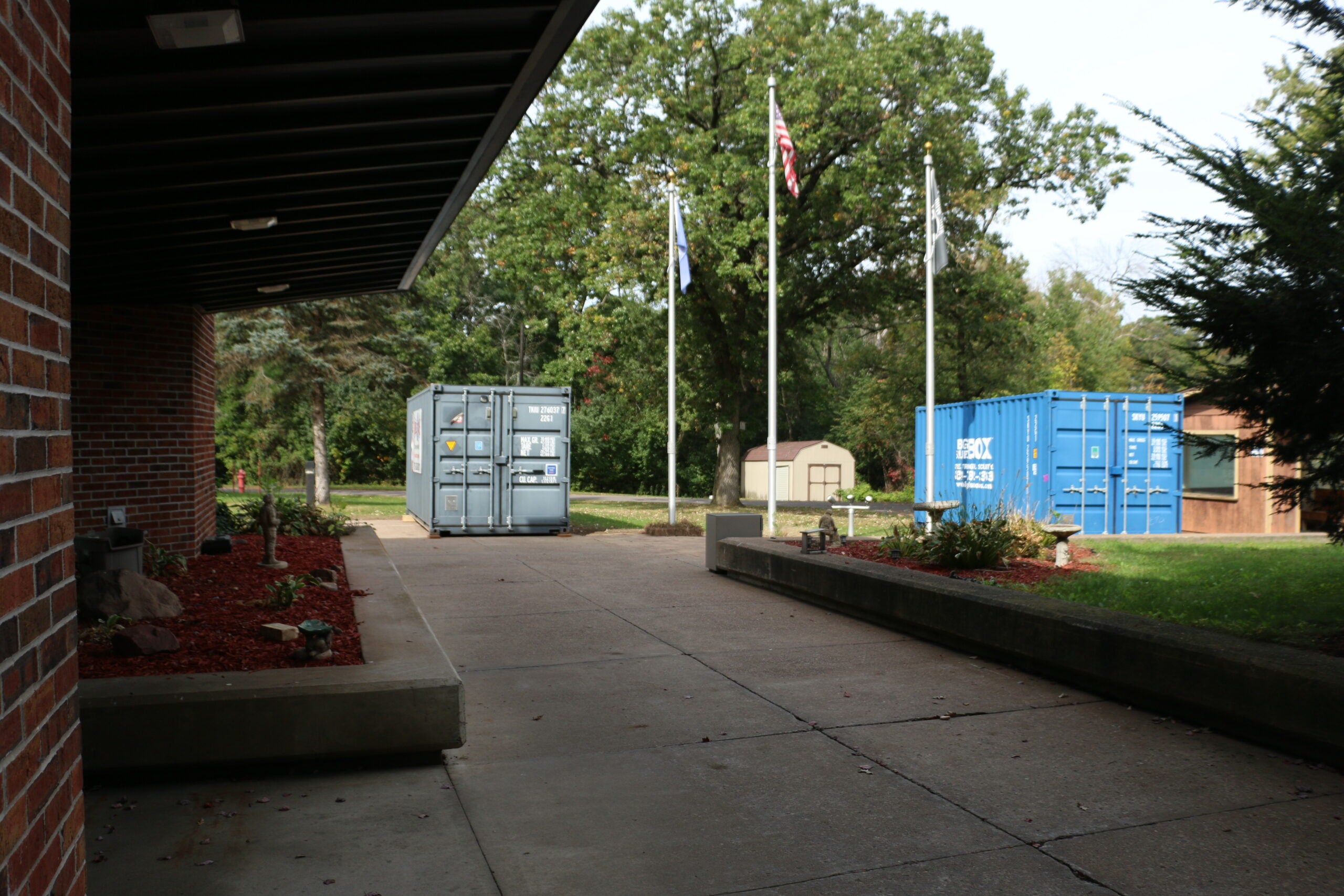 A concrete walkway between a brick building and landscaping, with two shipping containers and three flagpoles in the background. Trees and a small shed are visible.