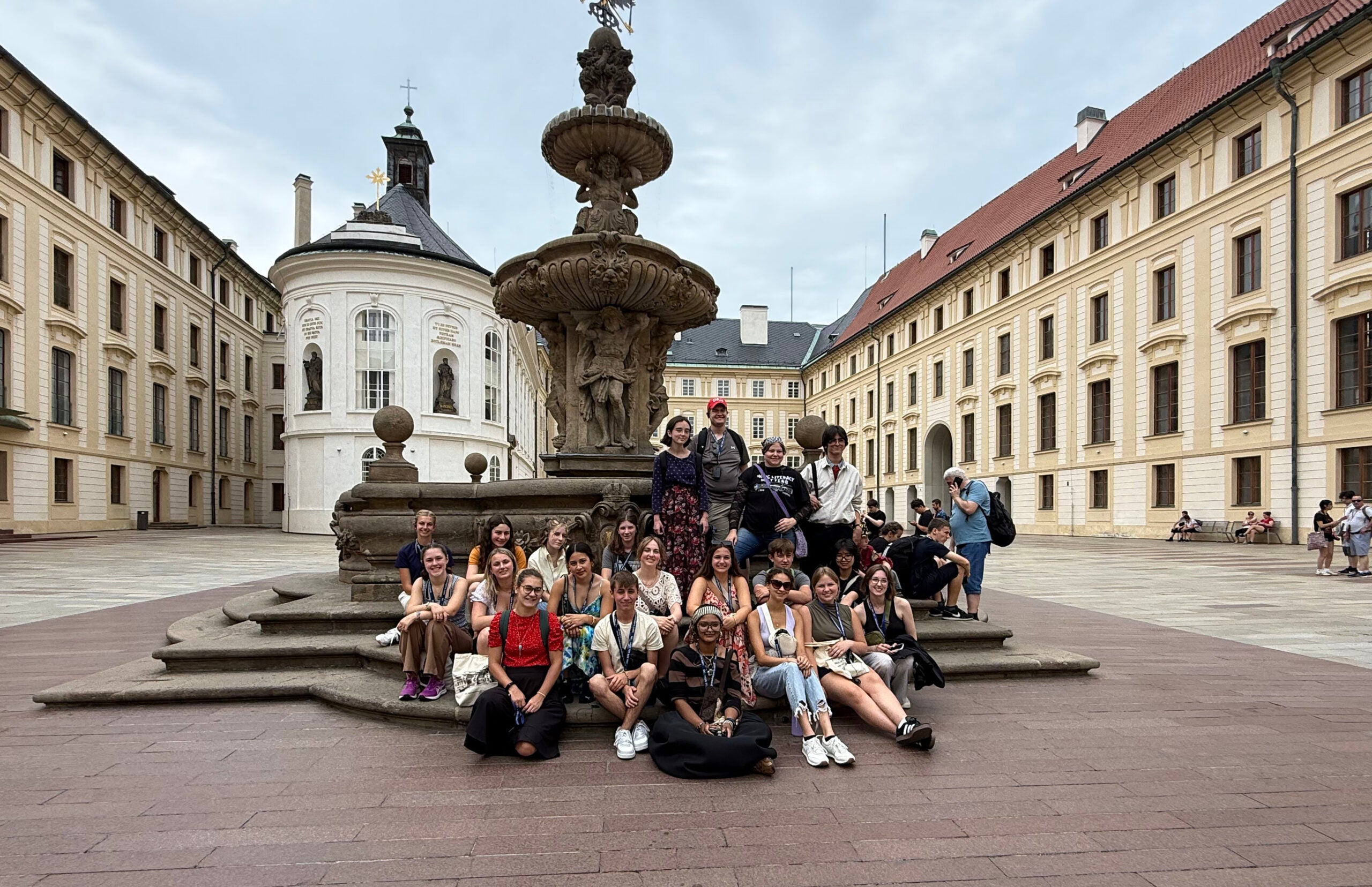 A large group of people pose in front of a stone fountain in the courtyard of a historic European building with arched windows and a chapel.
