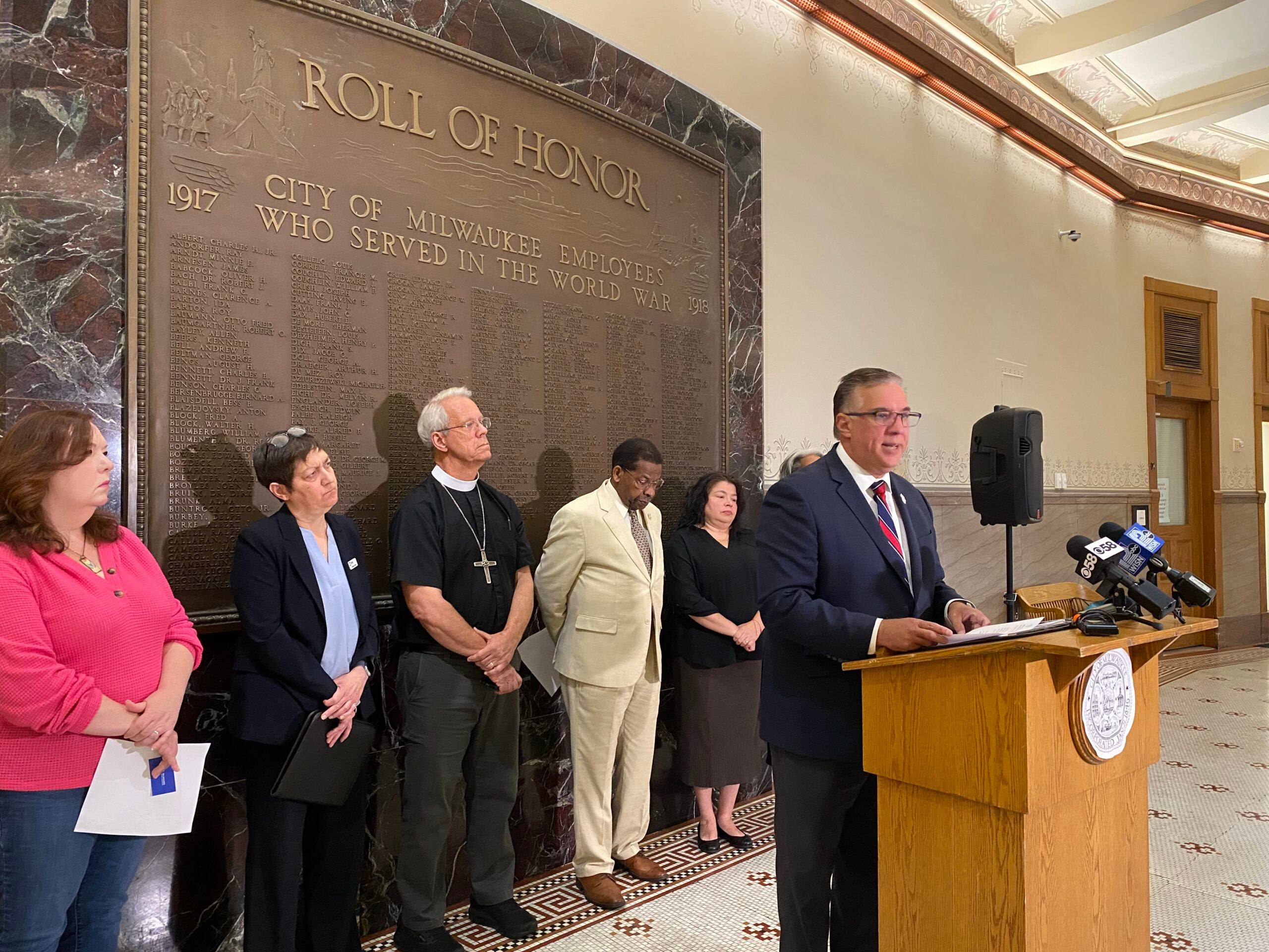 A man speaks at a podium in front of a wall memorial, with five people standing behind him, during a press conference in a government building.