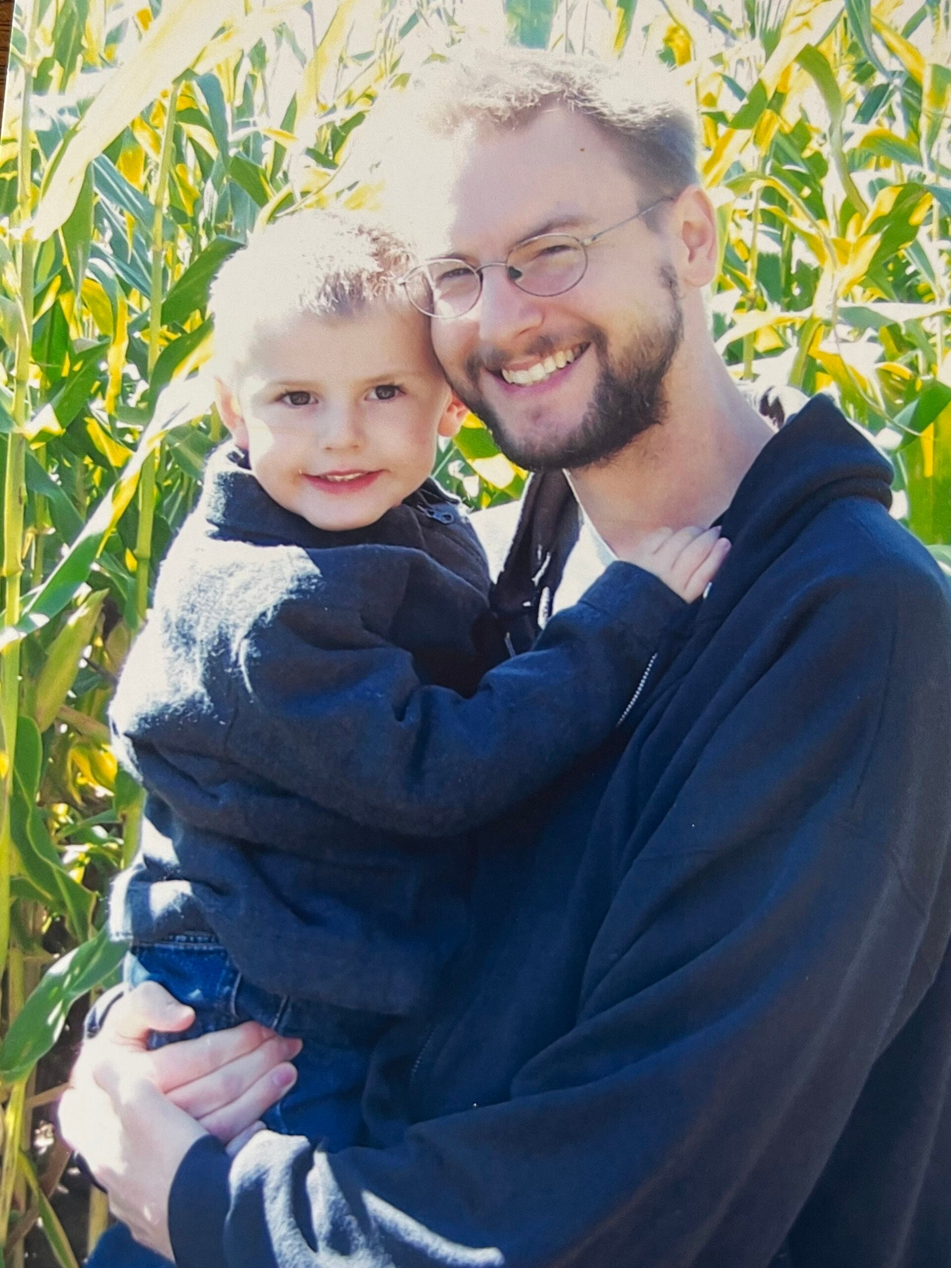 An adult and a child, both wearing dark jackets, smile while posing together in front of tall green corn stalks outdoors.