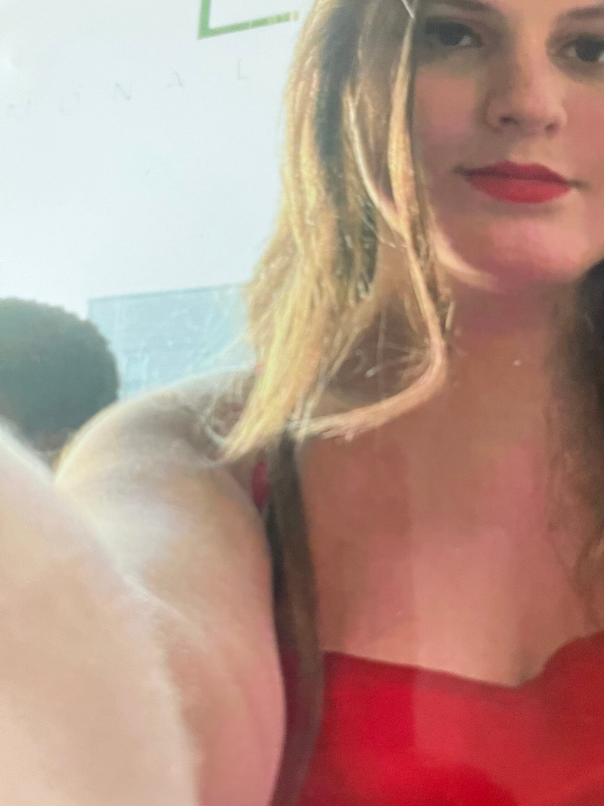 A person with long hair and red lipstick takes a selfie, wearing a red top, with part of their arm extending towards the camera. Another person is partially visible in the background.