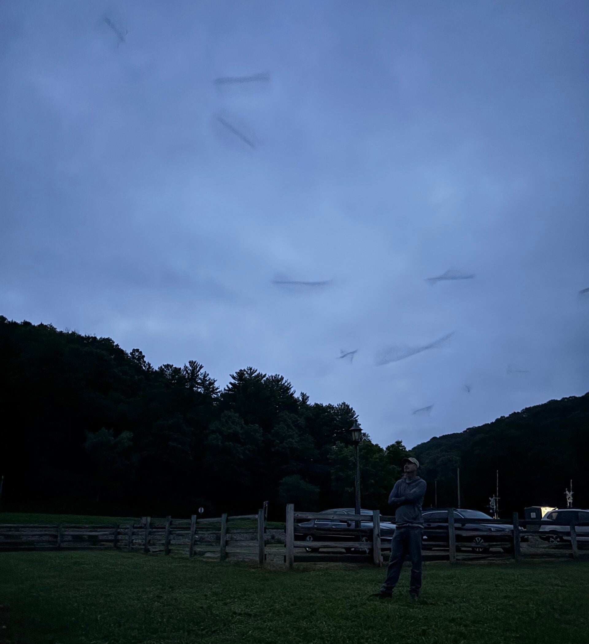 A person stands on grass near a wooden fence at dusk with several cars parked in the background and forested hills under a cloudy sky.
