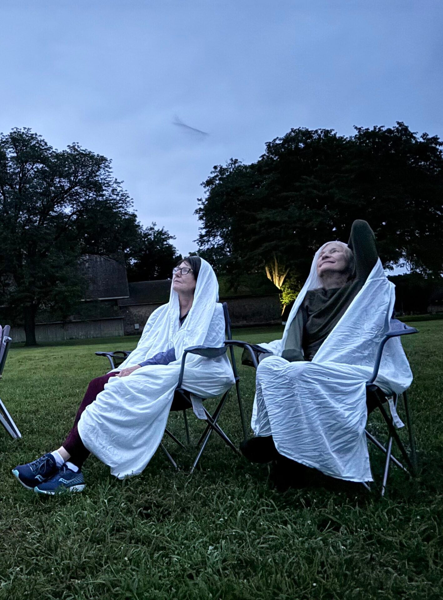 Five people sit on lawn chairs in a grassy field at dusk, wrapped in blankets, looking up at the sky. Trees and a building are visible in the background.