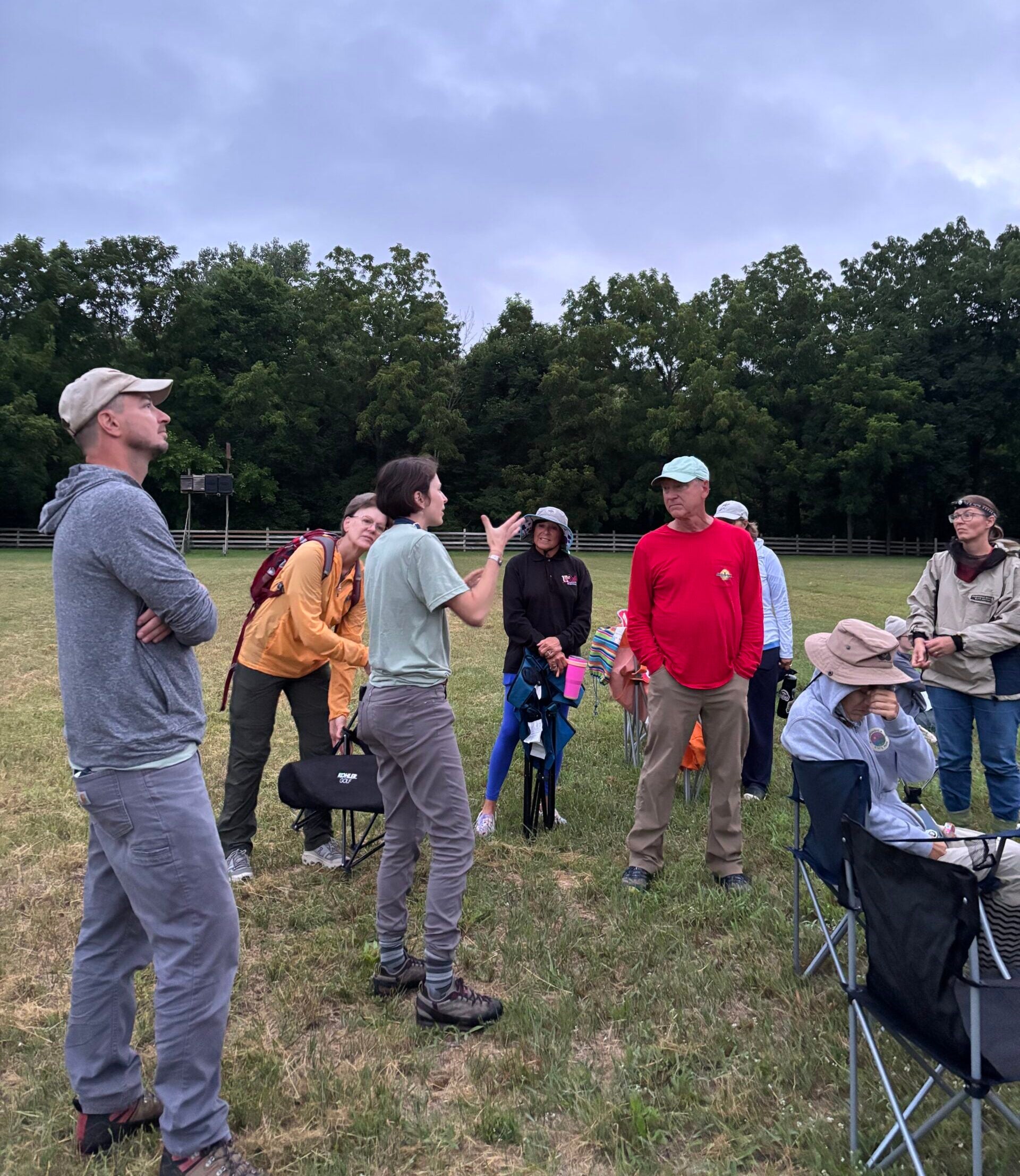 A group of people stand and sit in a grassy field, some listening to a person speaking while others sit in folding chairs. Trees are in the background under a cloudy sky.