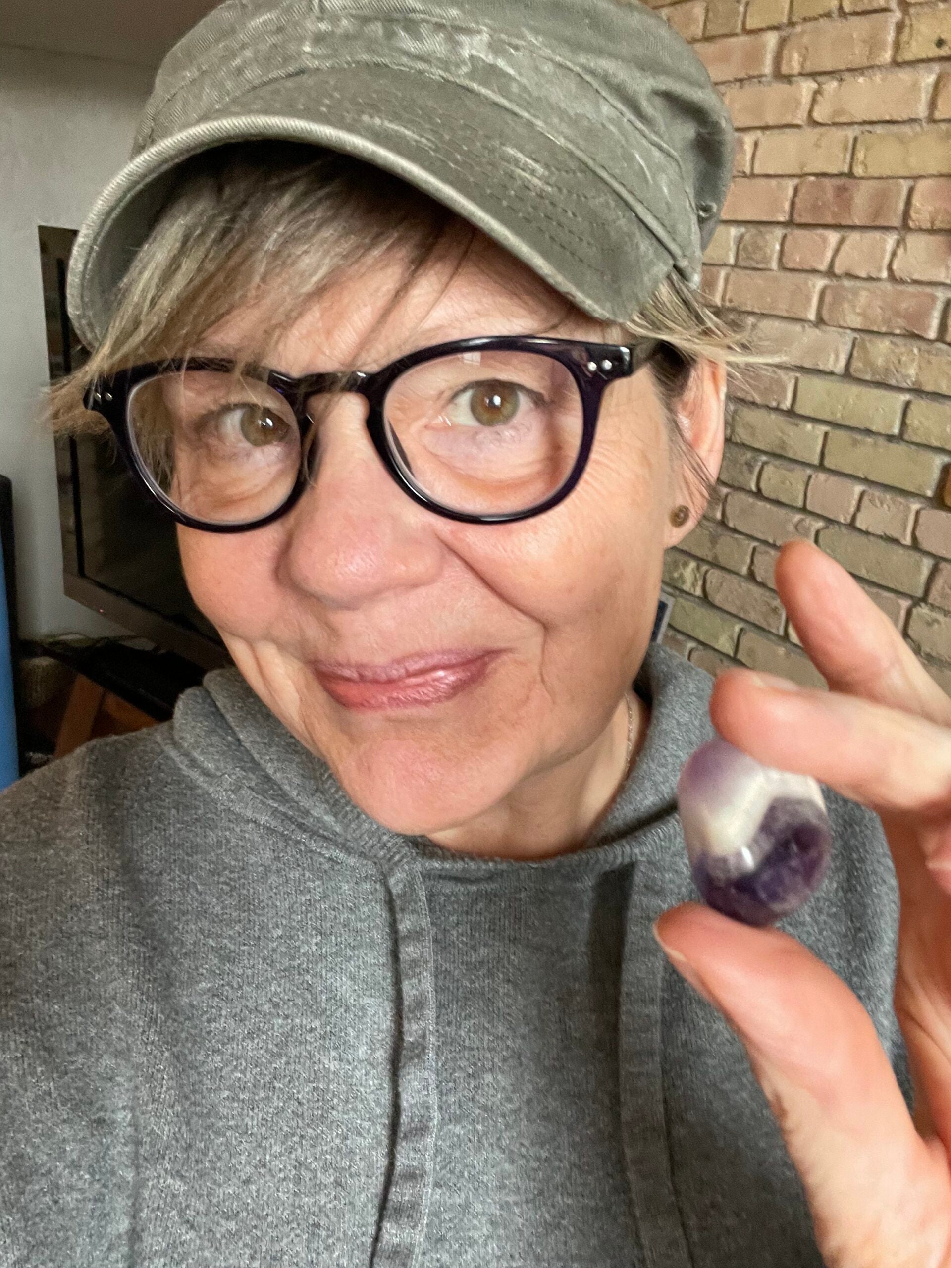 A person wearing glasses, a gray hoodie, and a cap holds up a small purple and white crystal, smiling at the camera.