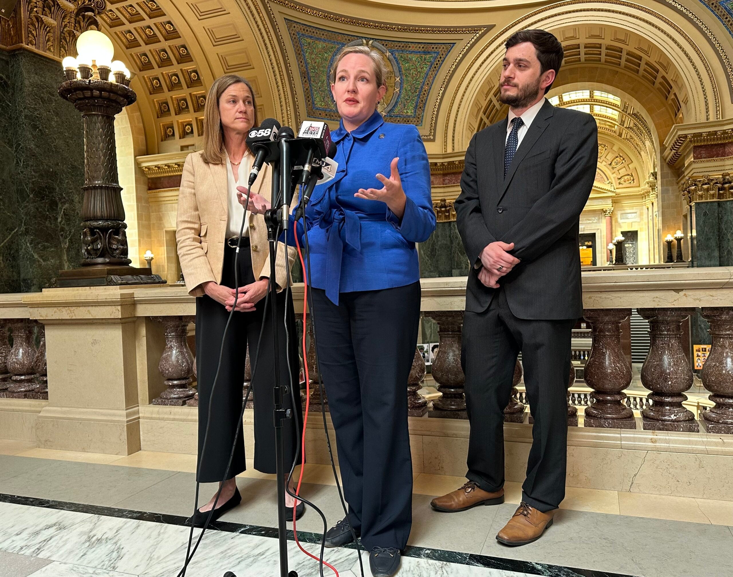 Three people in business attire stand indoors at microphones for a press conference, with ornate architectural details in the background.