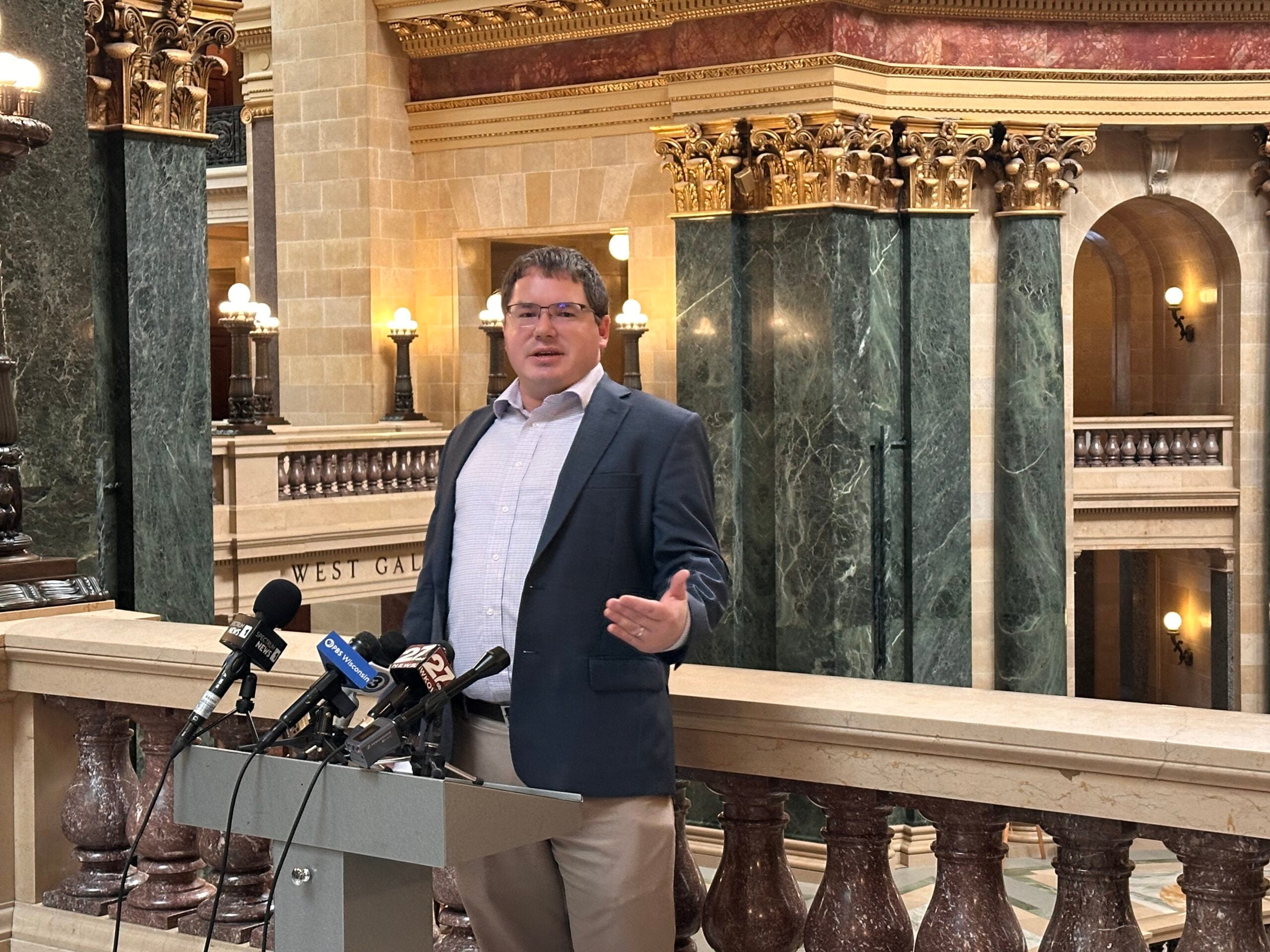 A man in business attire speaks at a podium with multiple microphones in a large, ornate government building.