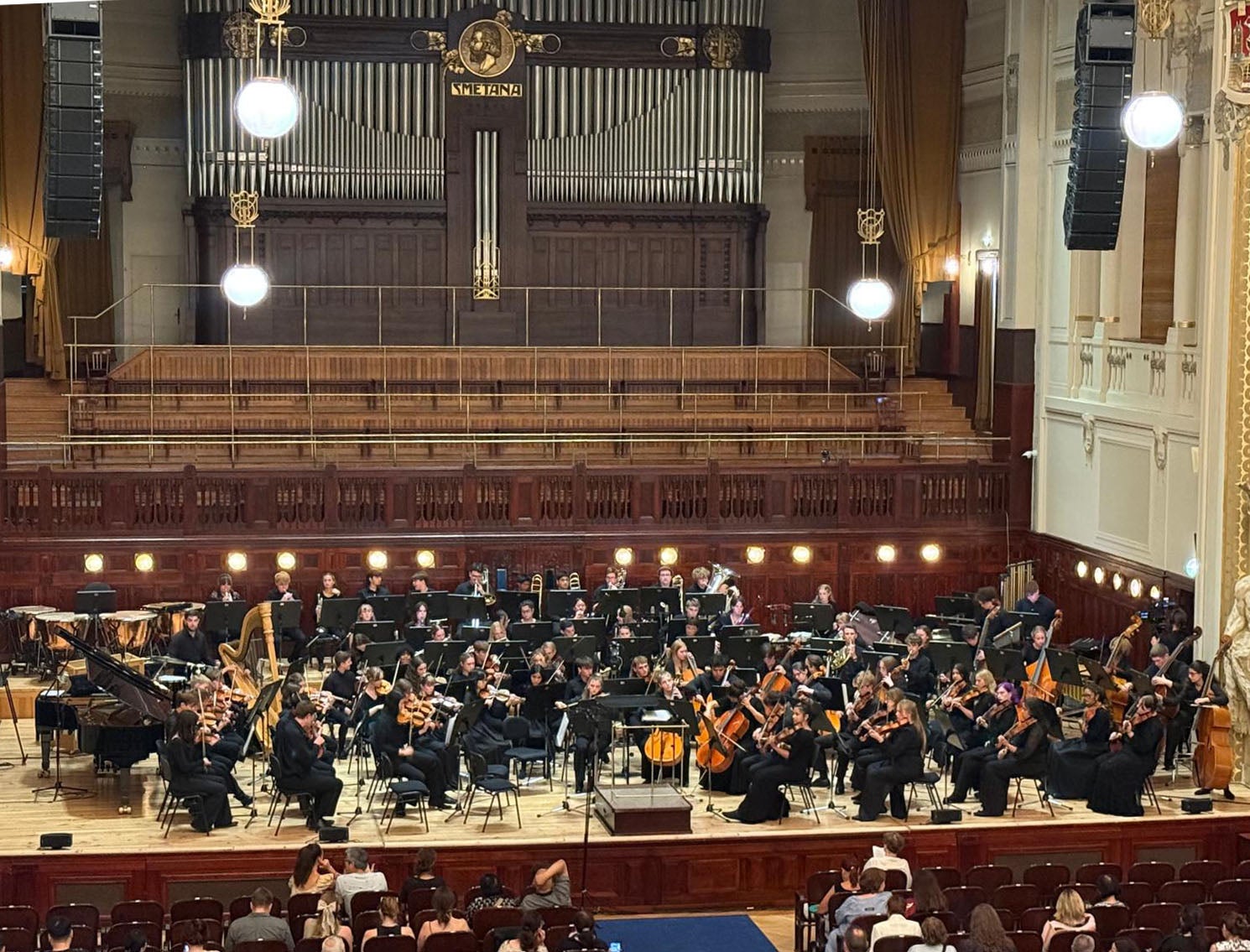 A full orchestra performs on stage in a grand concert hall with an organ in the background and an audience seated in front.
