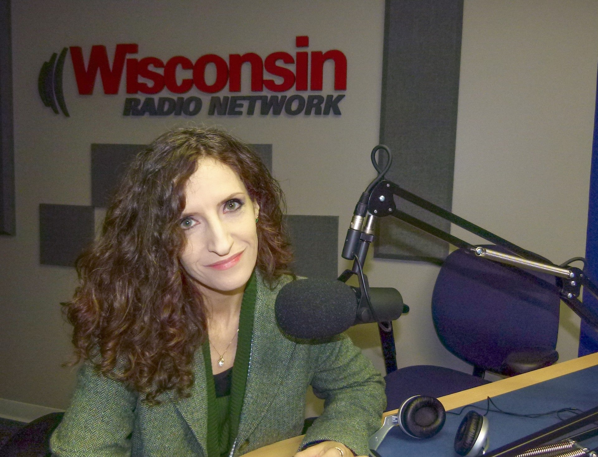 A woman with curly hair sits at a table with a microphone in a radio studio. The wall behind her has a Wisconsin Radio Network sign.