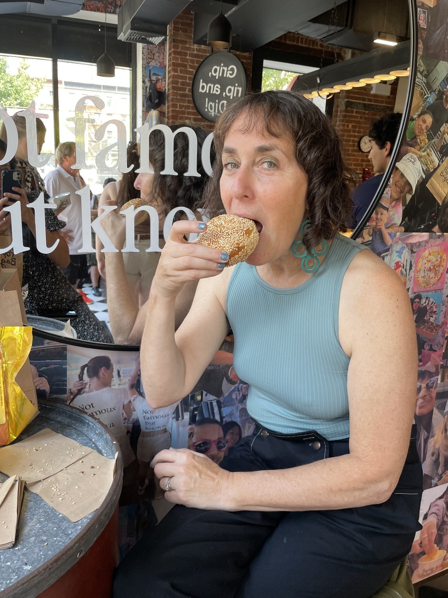 A woman in a sleeveless top sits at a café table, holding and eating a sesame seed bagel. The café has collage-covered walls and a large mirror.