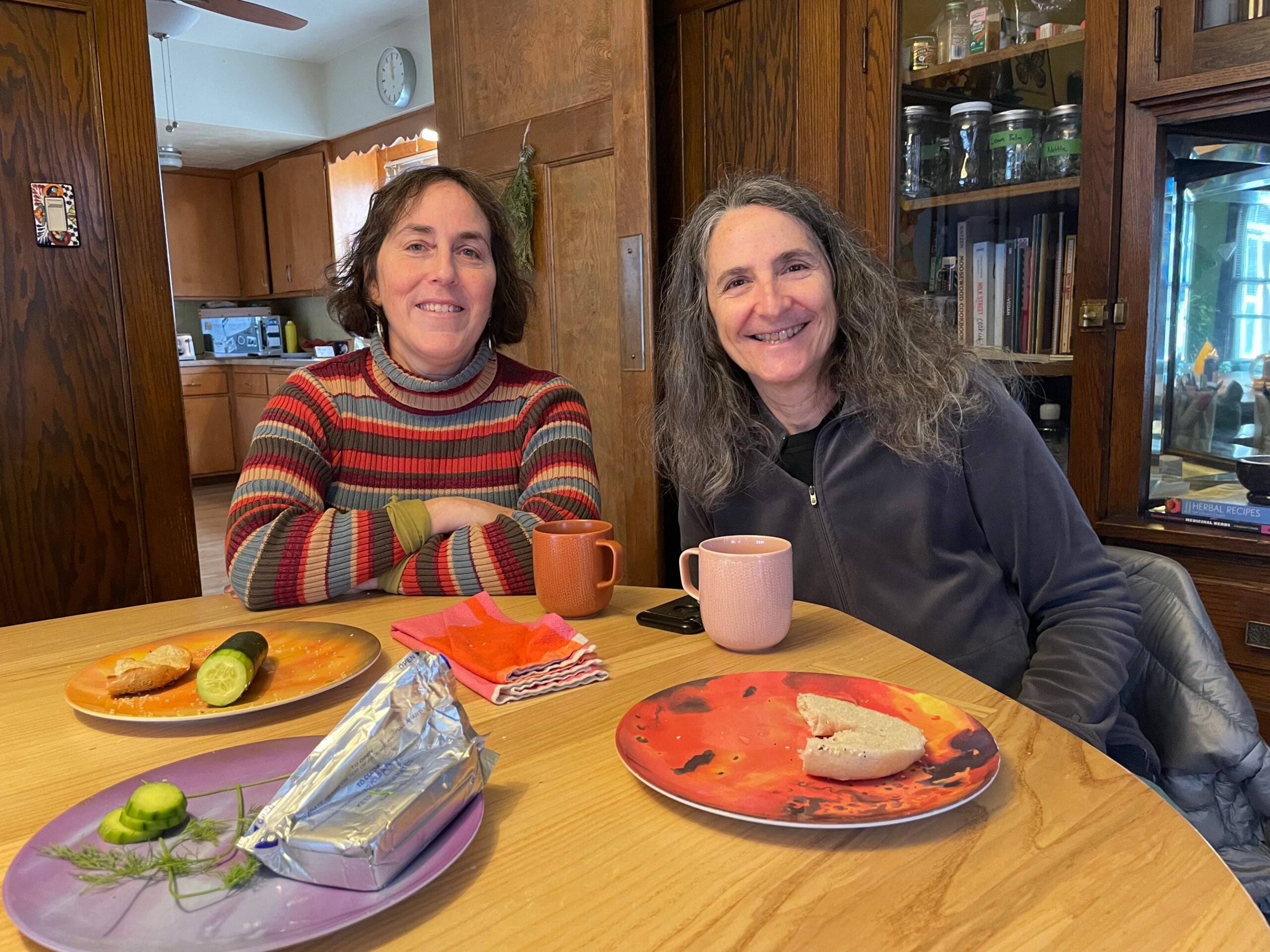 Two women sit at a wooden dining table with plates of food, mugs, and napkins in a cozy kitchen setting. Both are smiling at the camera.