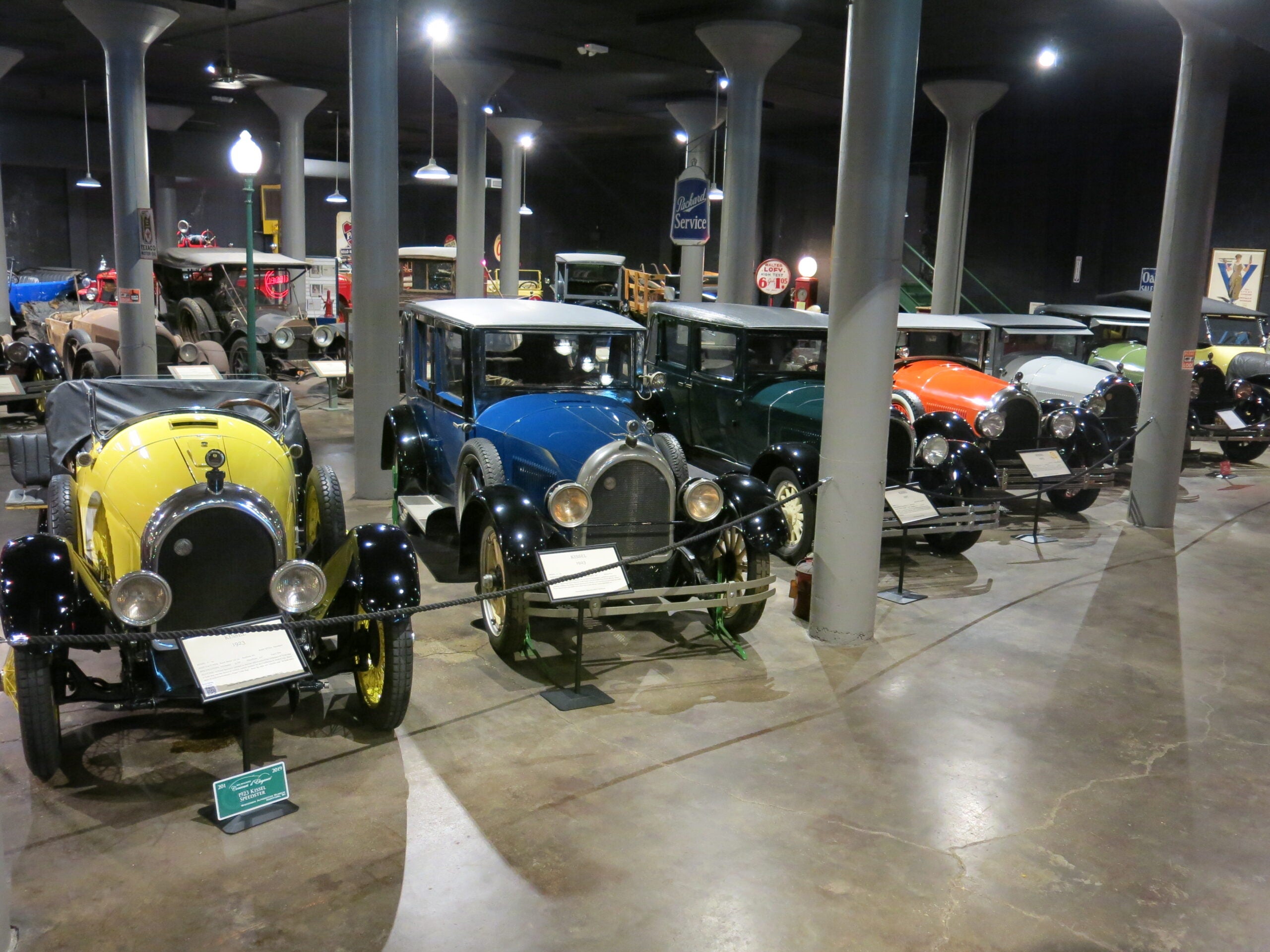 A row of vintage cars in various colors is displayed indoors at a museum, each with an informational sign in front.