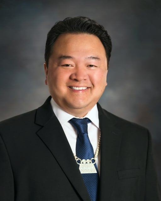 A man in a suit and tie is smiling, wearing a necklace with a rectangular pendant, posing against a dark, blurred background.
