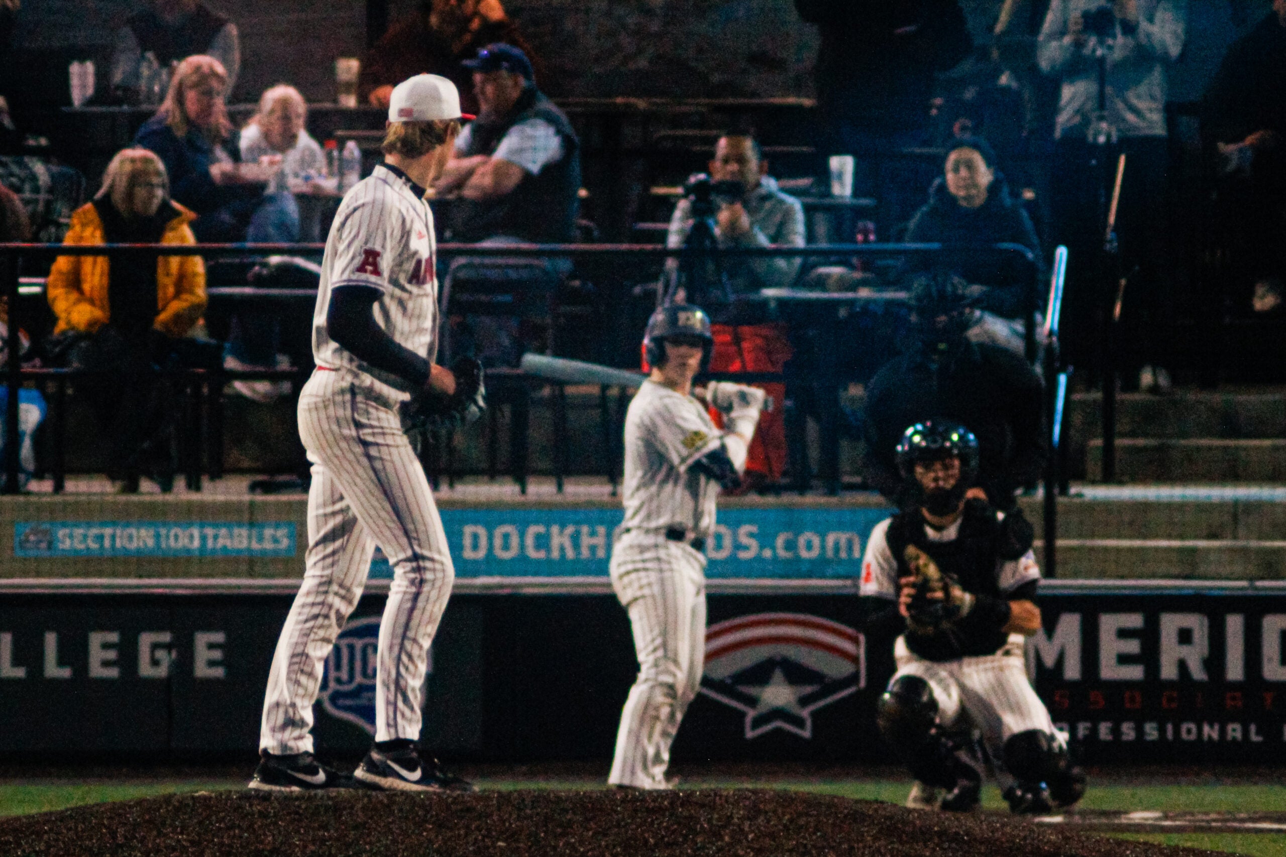 A baseball pitcher stands on the mound preparing to throw as the batter and catcher get ready, with spectators watching from the stands.