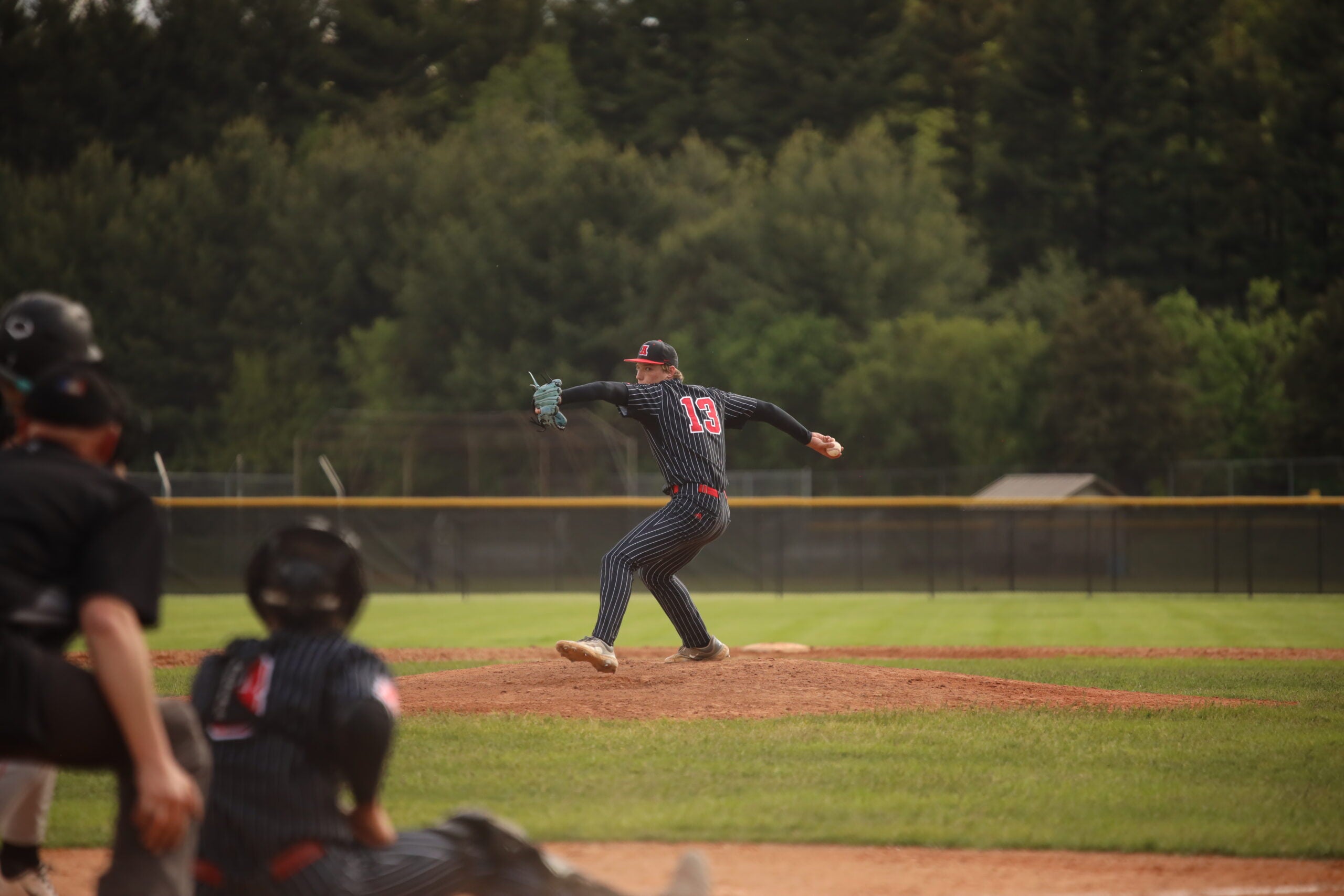 A baseball pitcher in a black and blue uniform prepares to throw the ball from the mound as a batter and catcher get ready for the pitch on a grassy field.