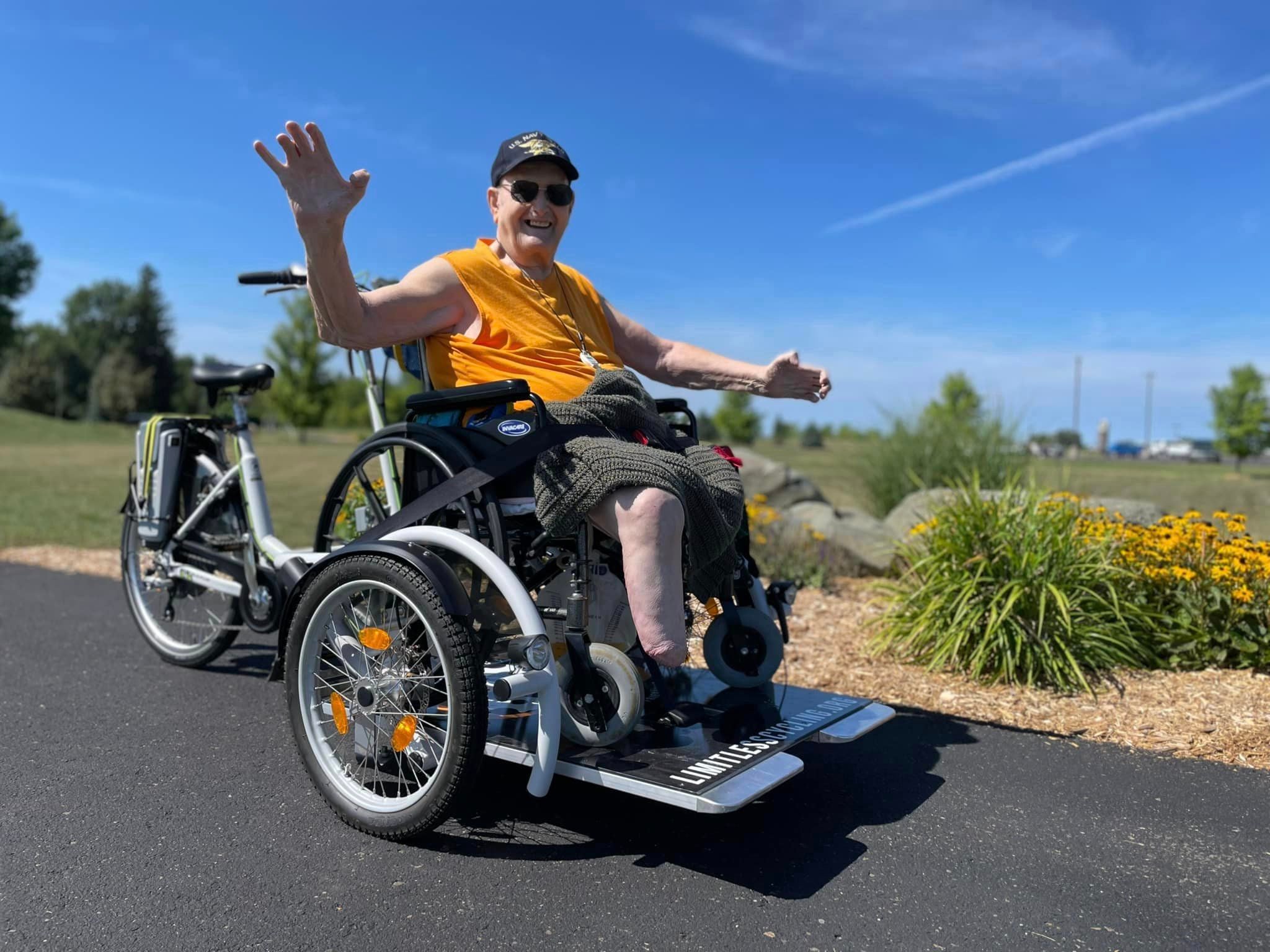 An older man in a wheelchair bike smiles and raises one hand while outdoors on a sunny day, with flowers and greenery in the background.