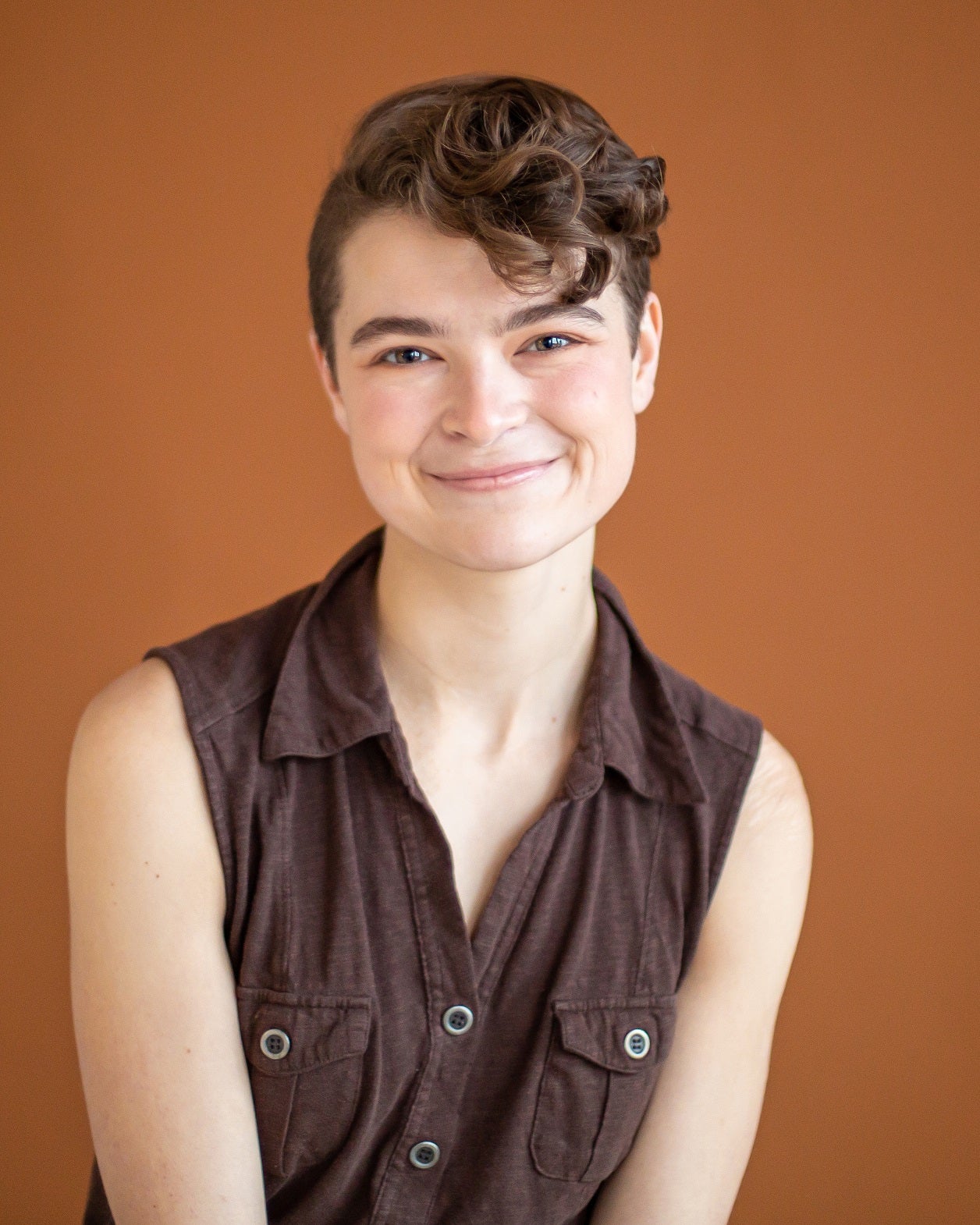 A person with short, curly hair and a sleeveless brown button-up shirt smiles in front of a solid orange background.