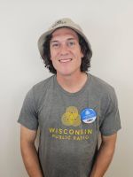 A person wearing a bucket hat and a Wisconsin Public Radio t-shirt stands against a plain background, smiling, with a blue sticker on their shirt.