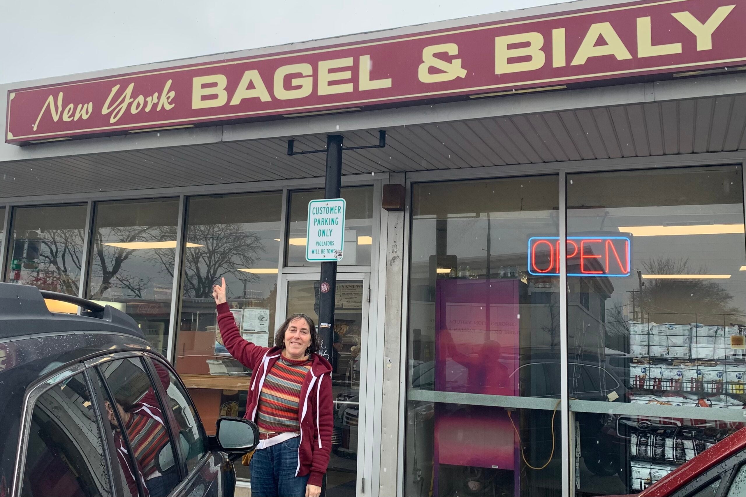A person stands outside New York Bagel & Bialy, pointing at the store sign. The storefront has an OPEN neon sign and a customer parking sign.