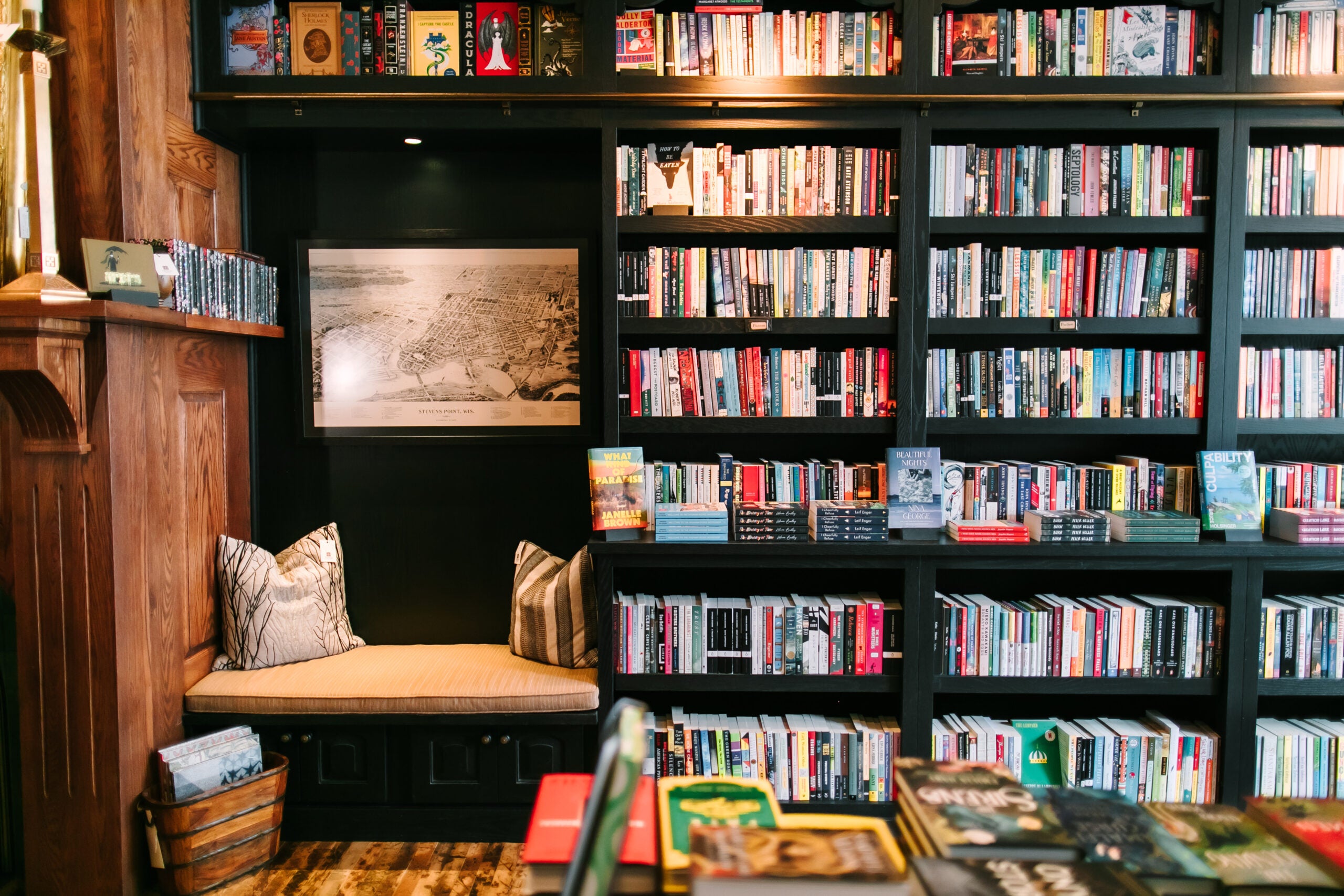 A cozy reading nook with cushions beside black bookshelves filled with books in a well-lit library or bookstore.