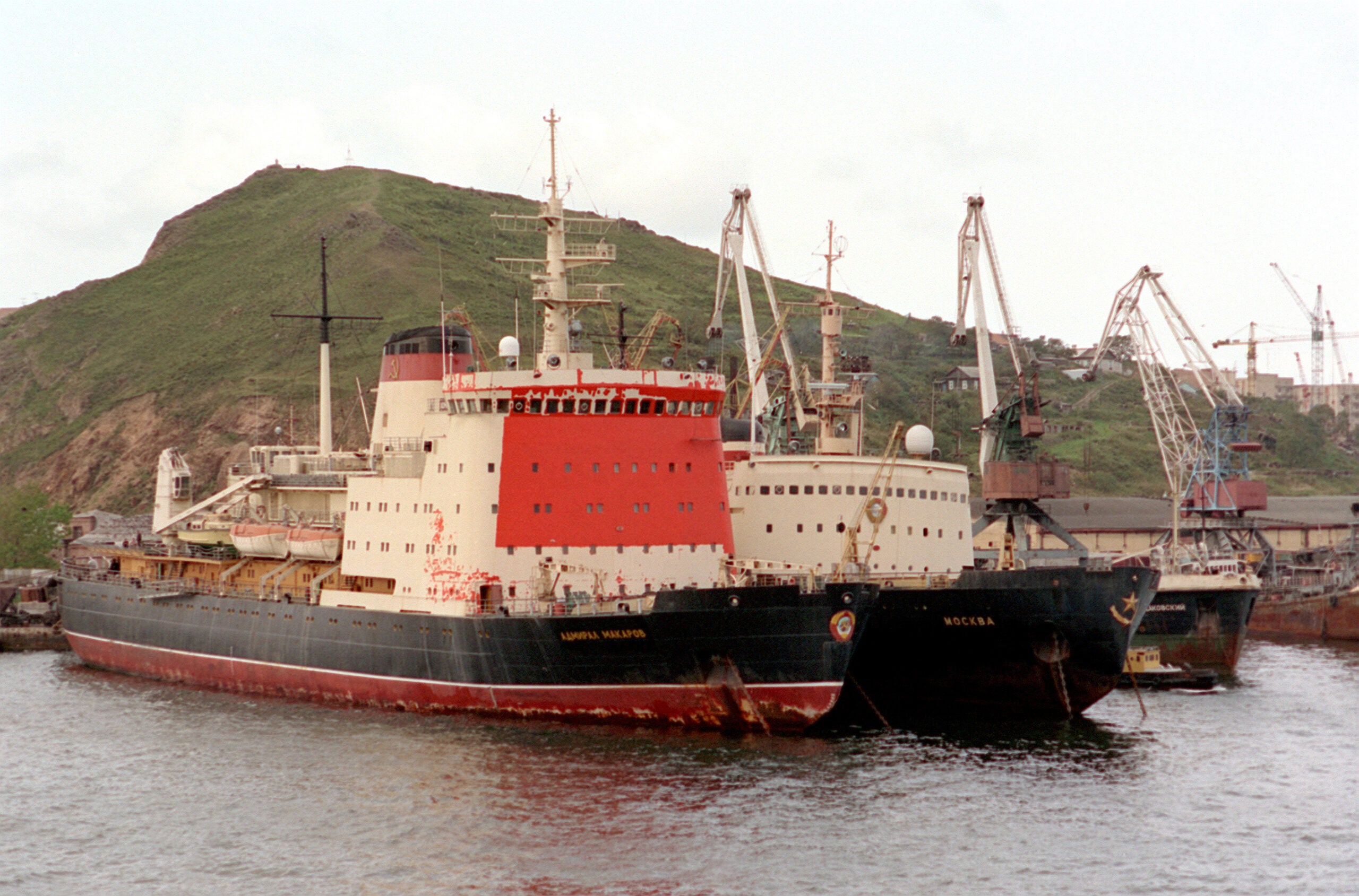Two large icebreaker ships are docked side by side at a port, with cranes and a green hill visible in the background.