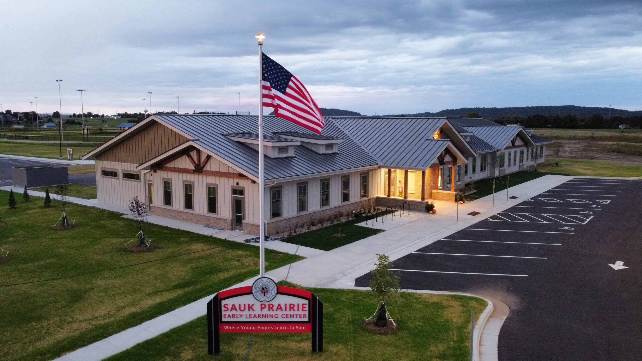 A large building with a metal roof houses the Sauk Prairie Early Learning Center, with an American flag and sign out front and an adjacent parking lot.