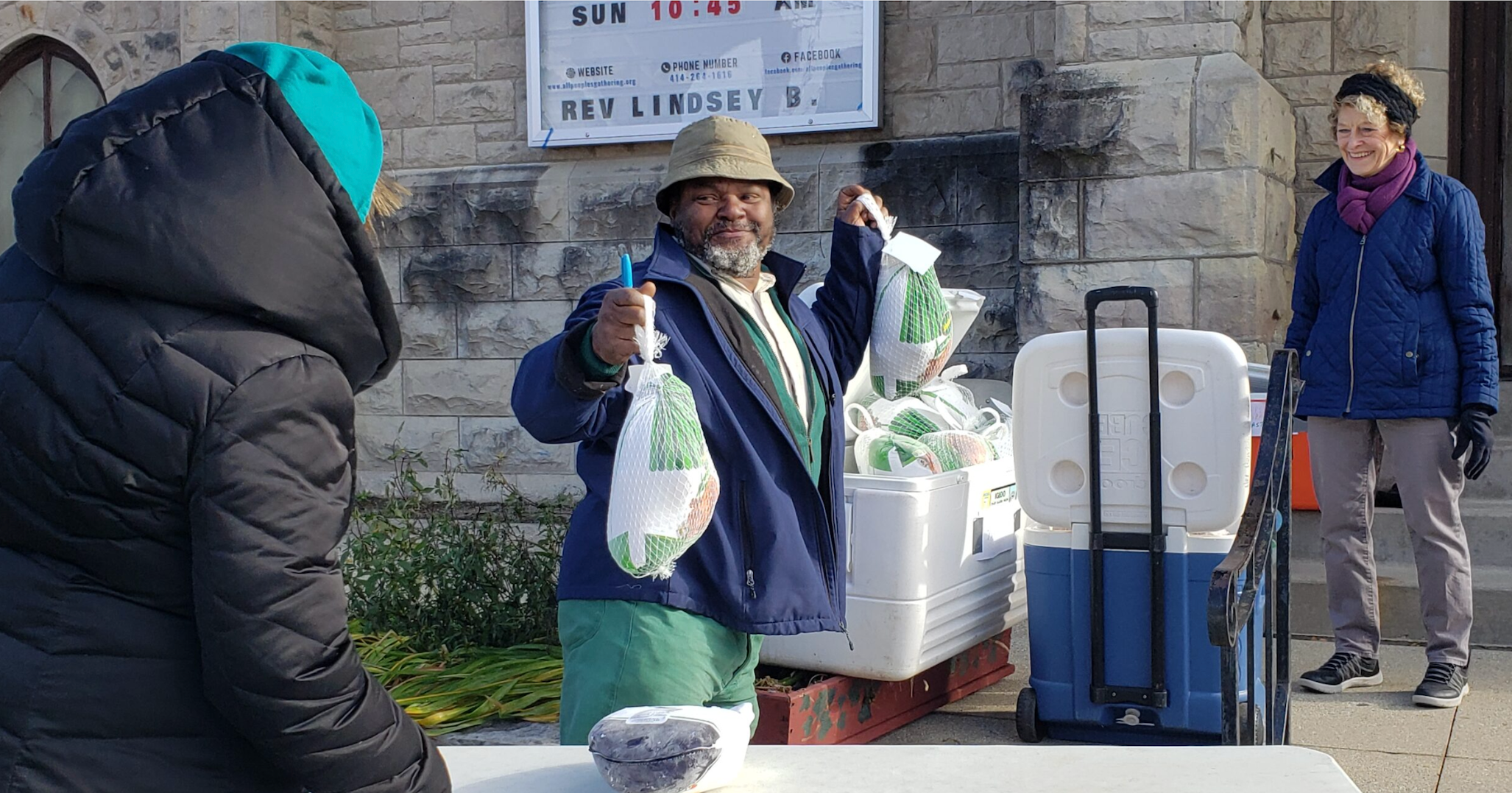 A man stands outdoors holding up two frozen turkeys, while two women stand nearby; a cooler and more turkeys are on a table in front of a stone building.